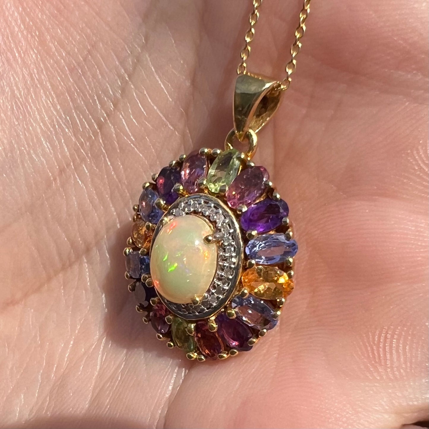 A gold plated Ethiopian fire opal and multi-gemstone halo pendant shown from an angle in sunlight.