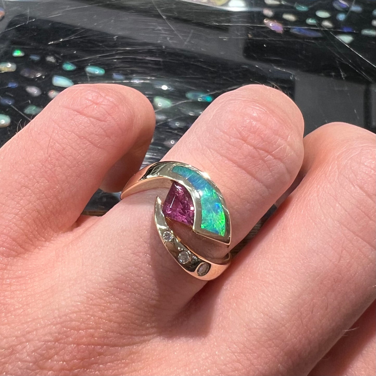 A 14k gold twisted shank ring set with a triangle cut pink tourmaline, green semi-black opal inlay, and three flush-set diamonds.