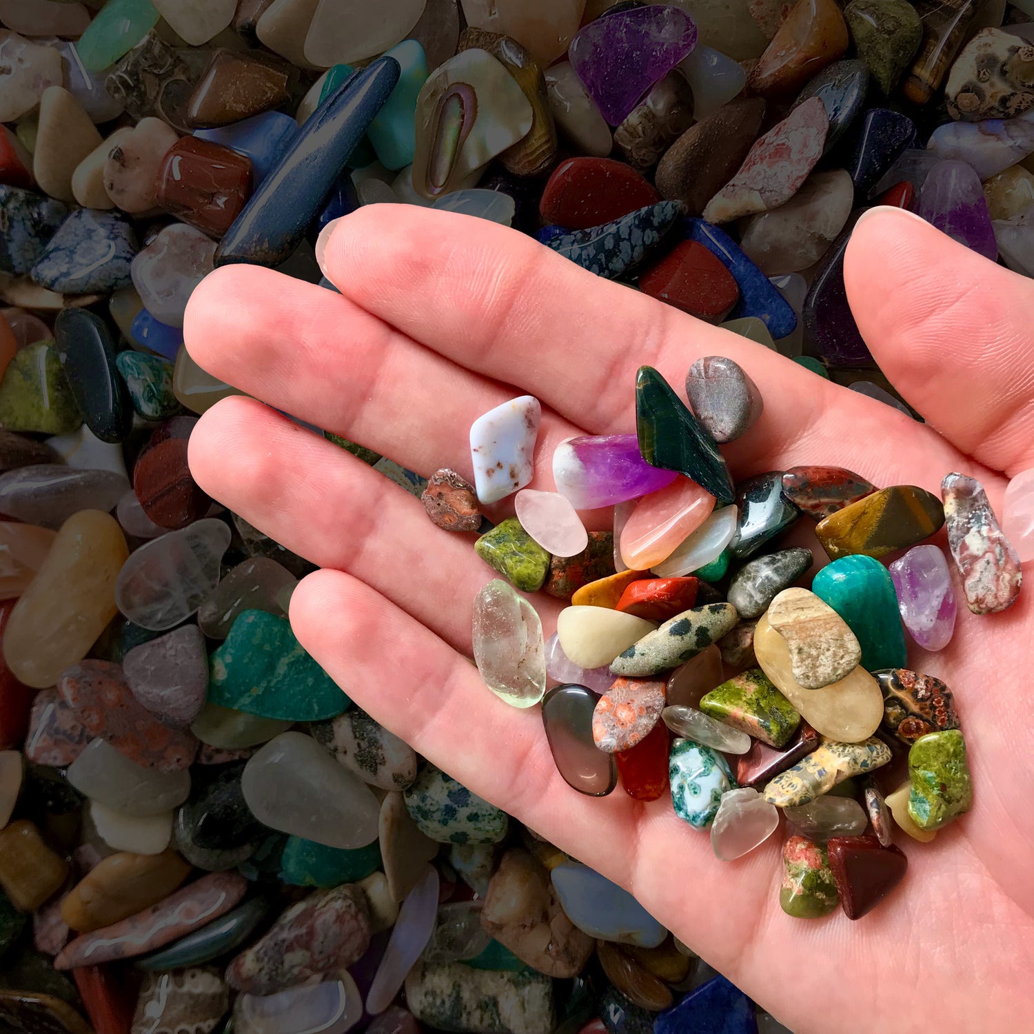 A hand holding a spray of colorful tumbled gemstone chips. Minerals include unakite, amethyst, and polished crystals.