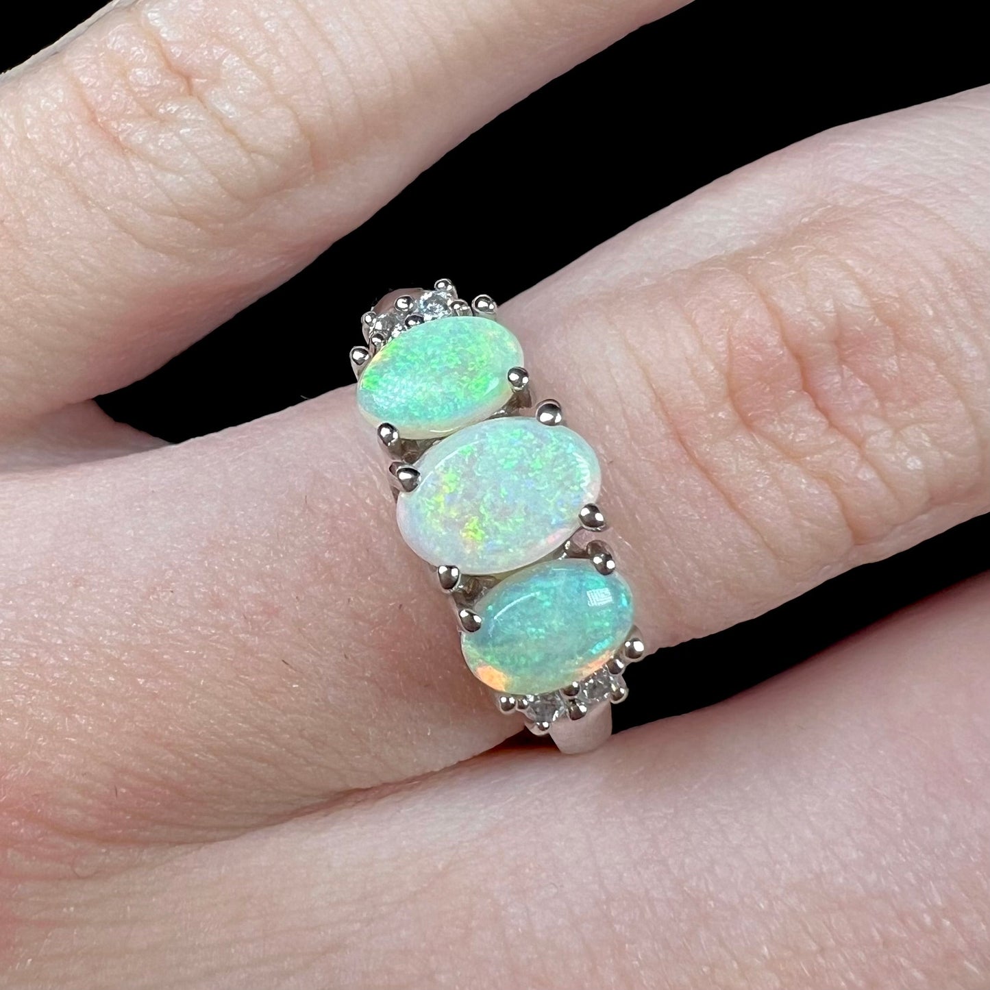 A ladies' white gold ring prong-set with three natural white opals and diamond accents. The opals play a green color.
