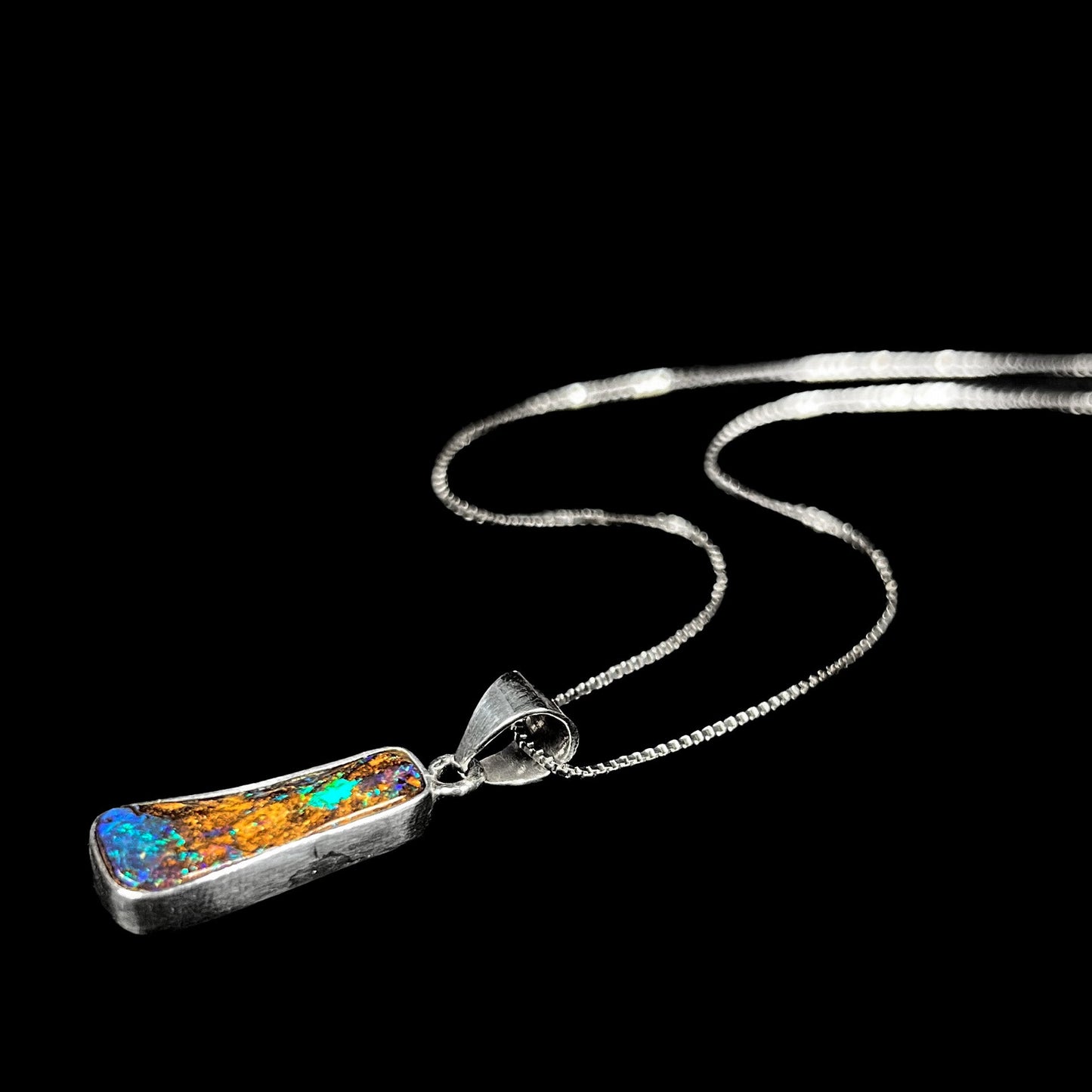 A sterling silver necklace bezel set with a natural Australian boulder opal stone. The stone shines colors of blue, green, and purple.