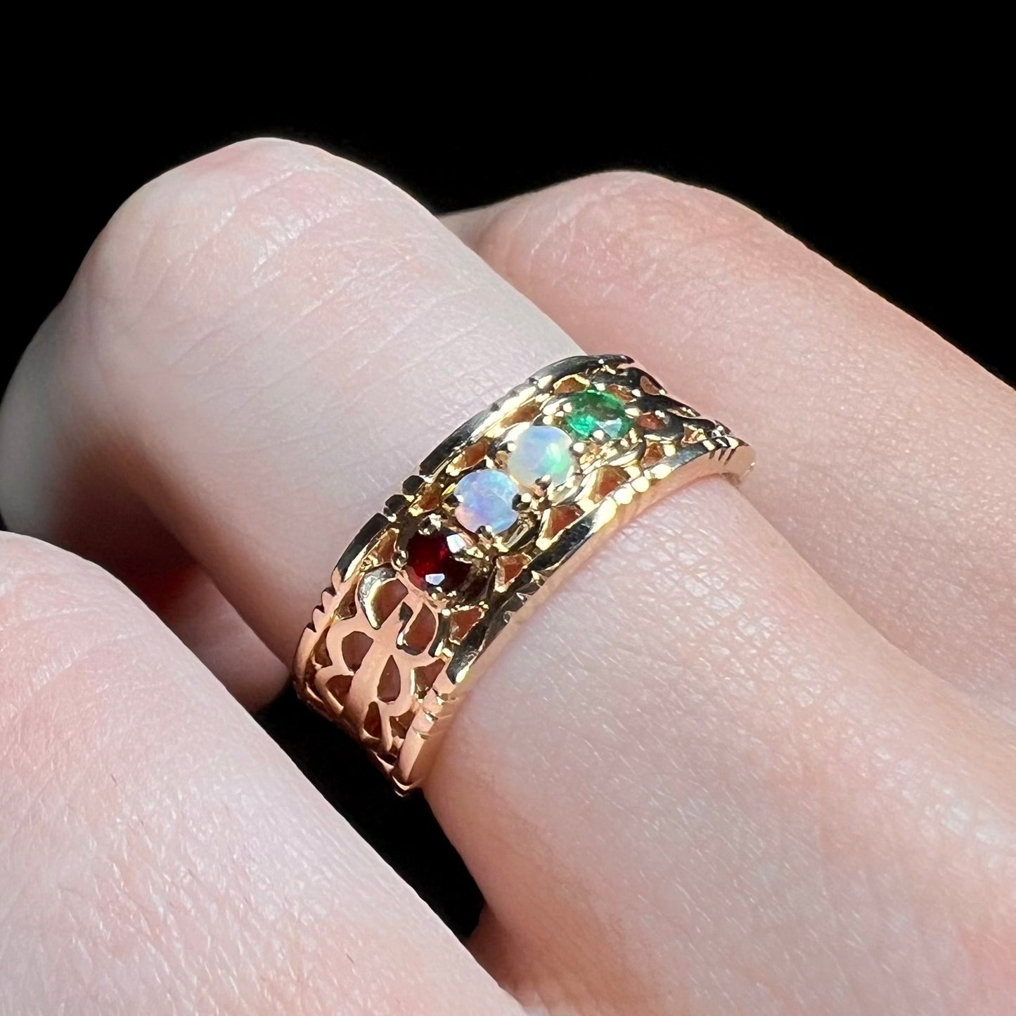 A gold woven-style mother's ring set with two opals, an emerald, and a garnet worn on a finger, shown at an angle.
