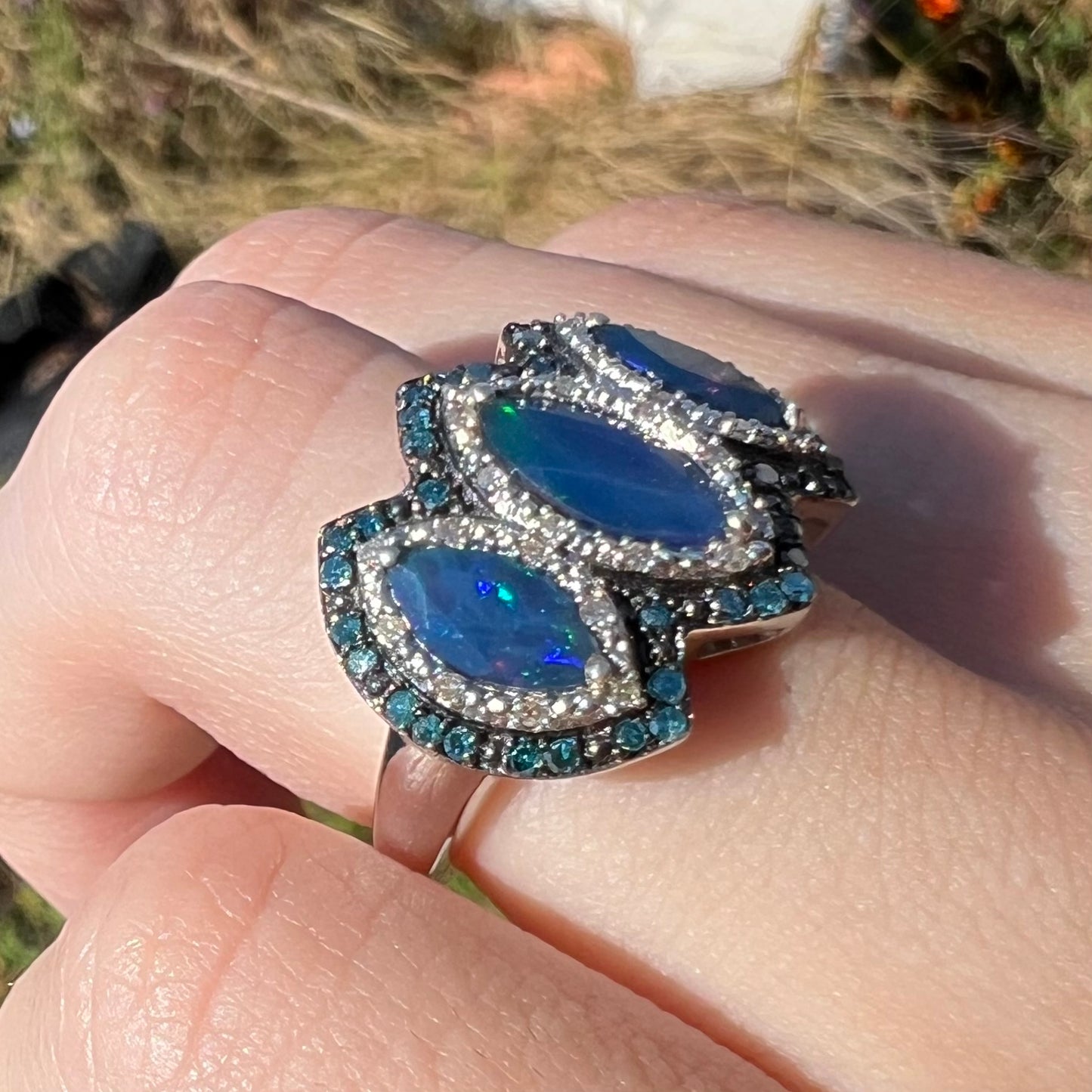 A three-stone opal and blue diamond ring worn on a finger at an angle in natural light.