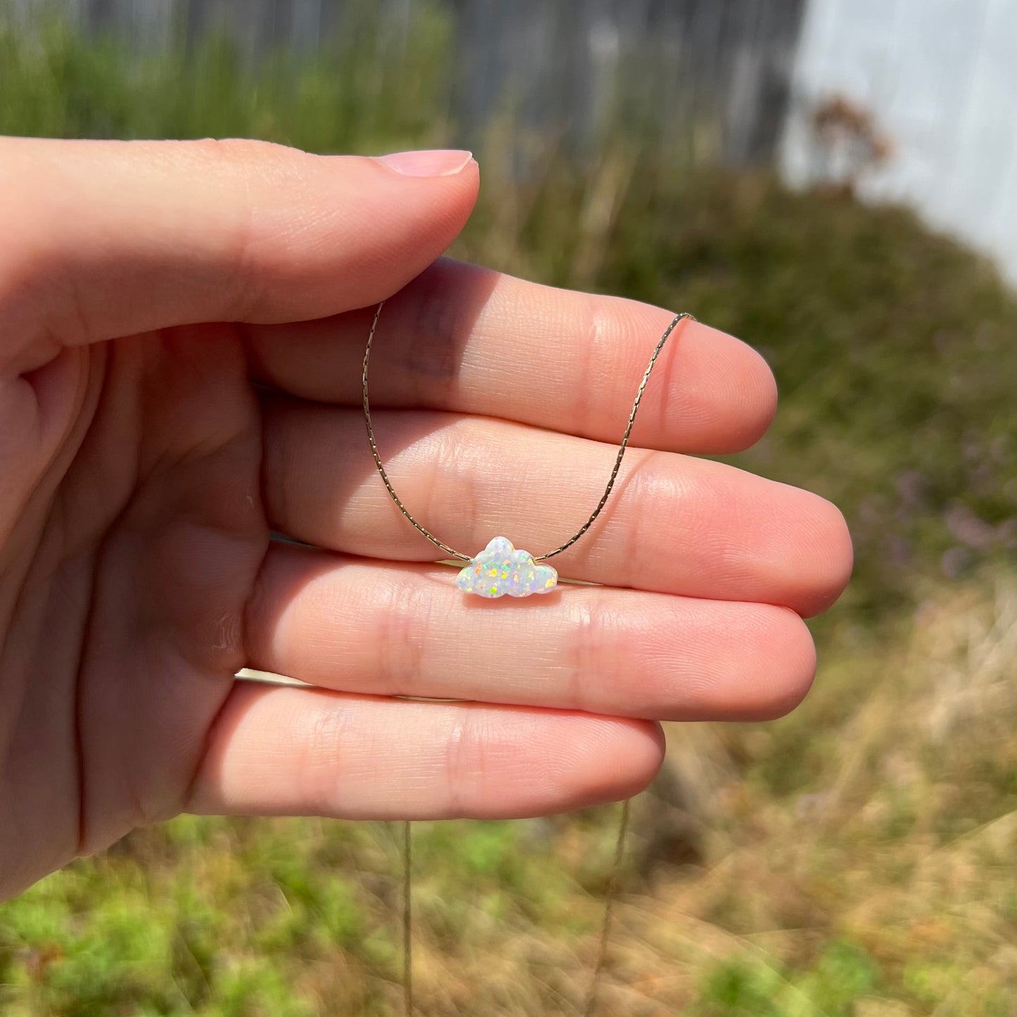 A lab-created opal cloud necklace on a gold filled chain held in a hand at a distance in the sunlight.