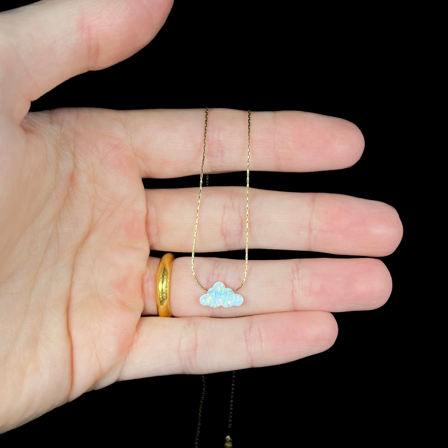 A lab-created opal cloud necklace on a gold filled chain held in a hand against a black background.