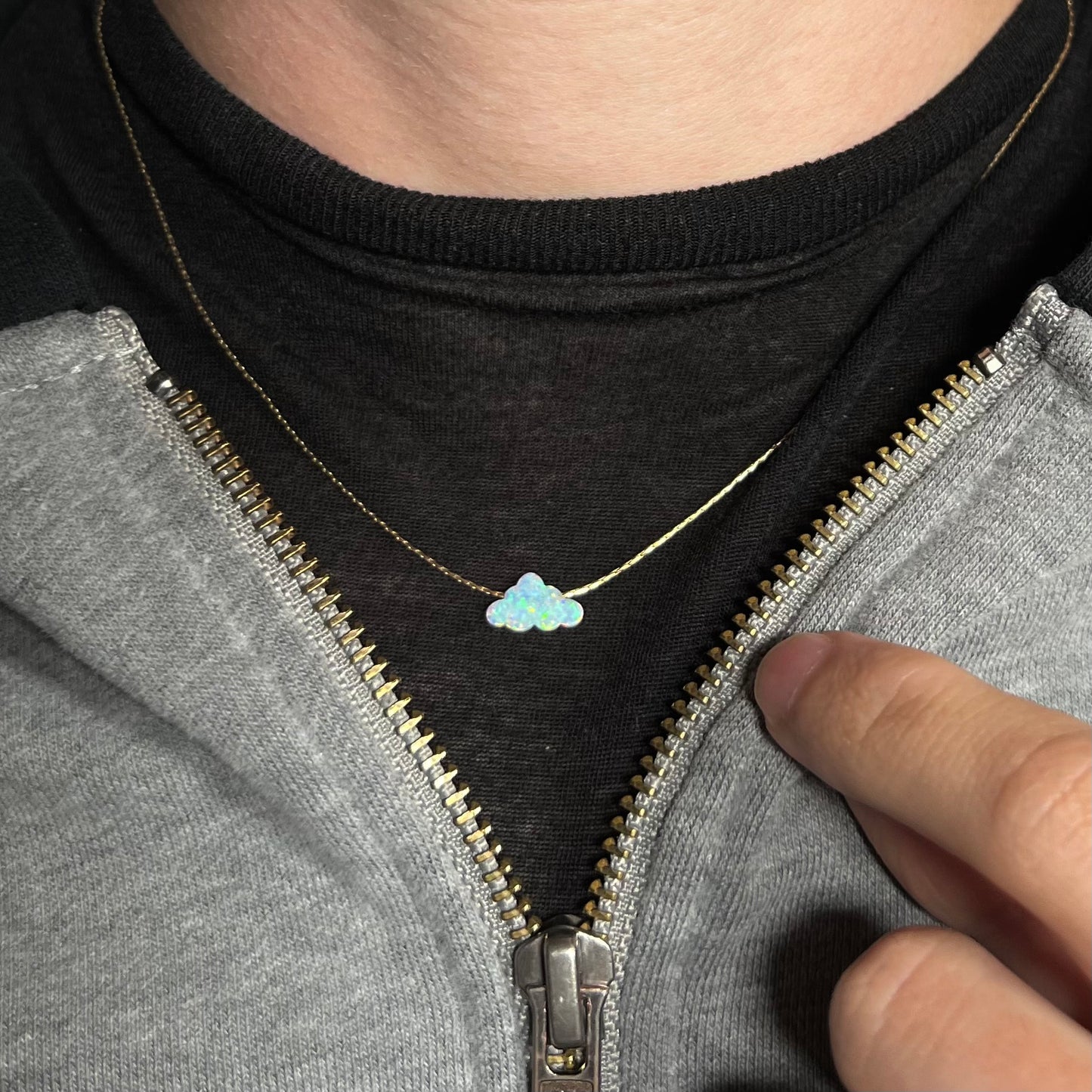 A lab-created opal cloud necklace on a gold filled chain worn around on a dark shirt under a partially unzipped hoodie.