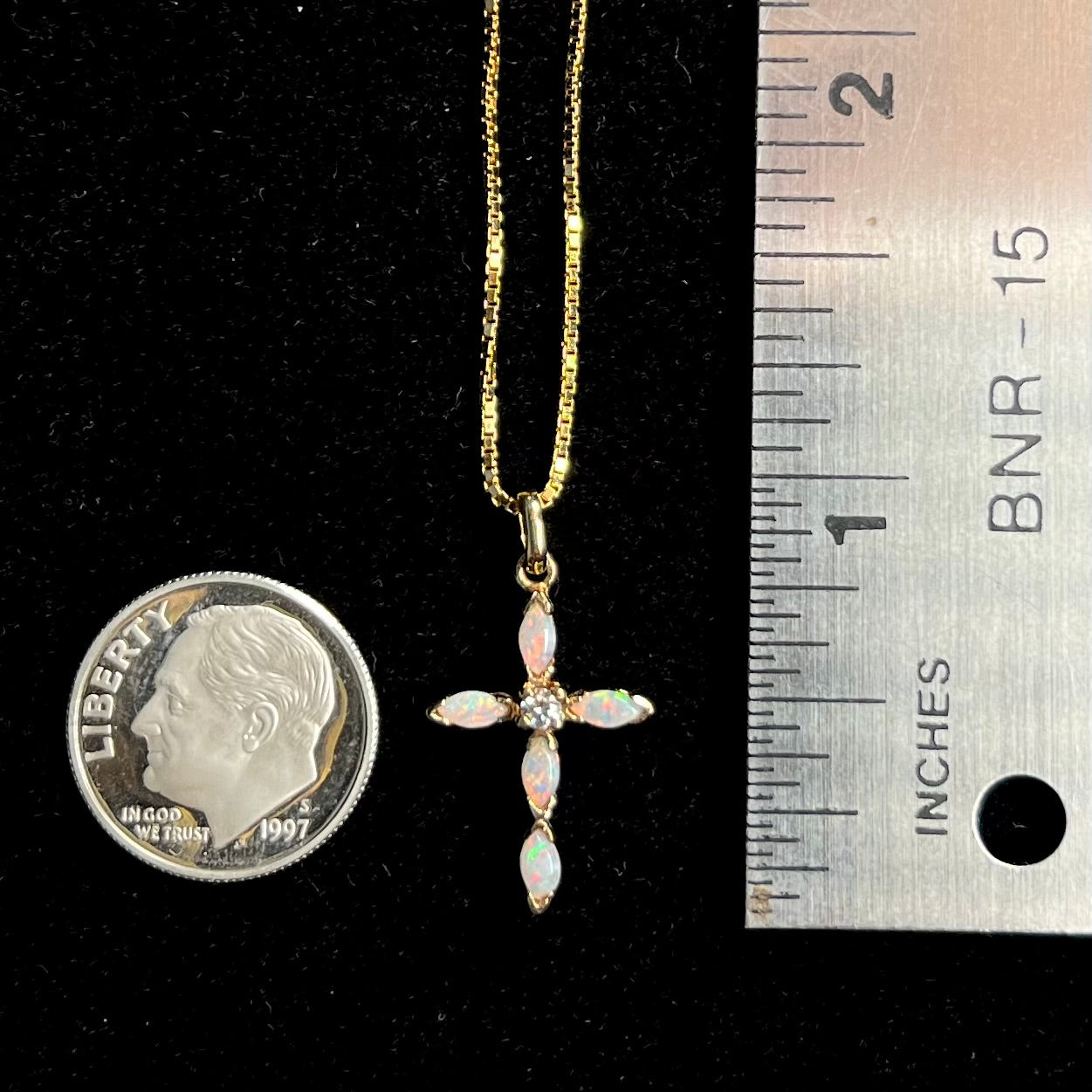 An opal cross necklace next to a dime coin and ruler for scale.