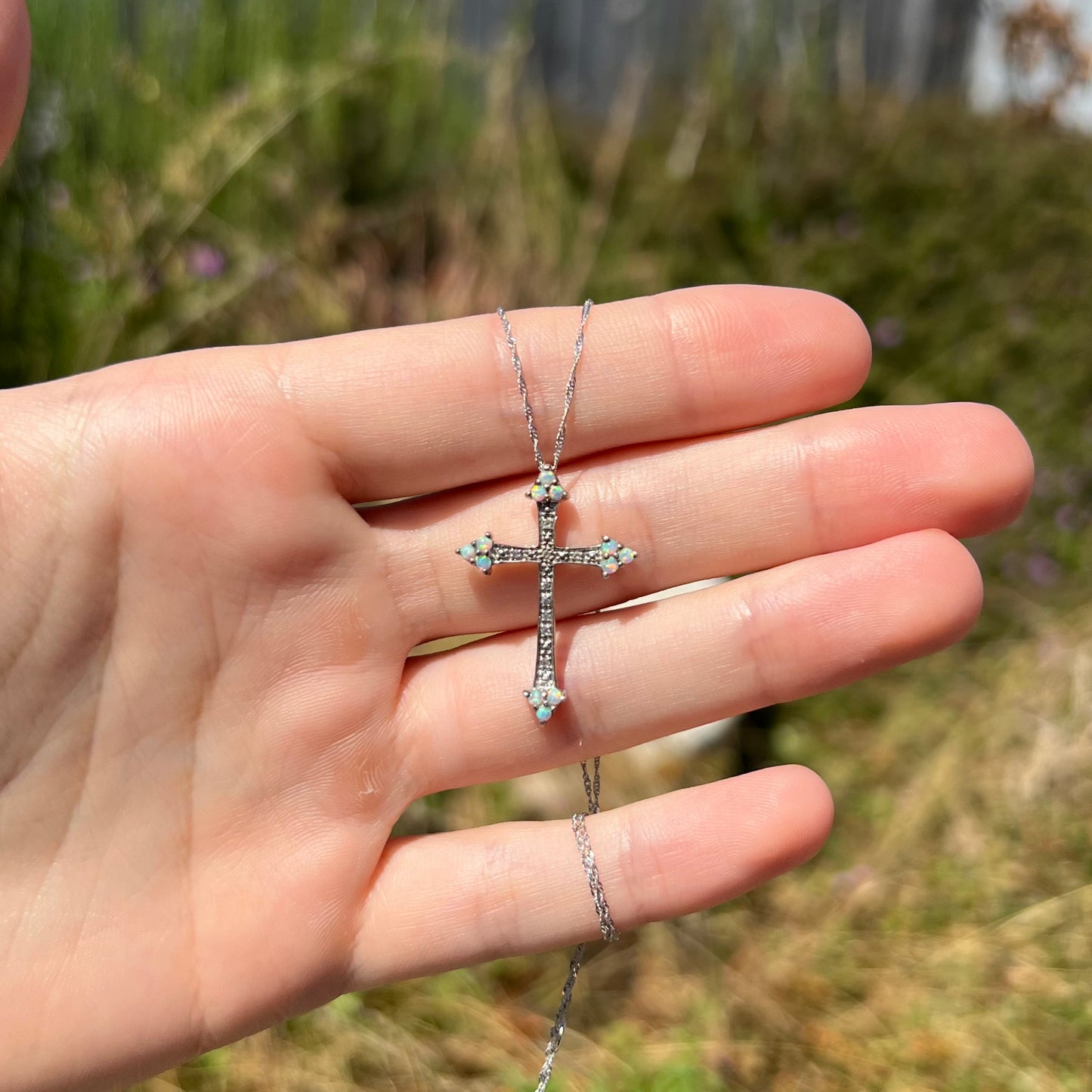 A sterling silver lab-created opal cross necklace with CZ accents held in a hand in sunlight at a distance.