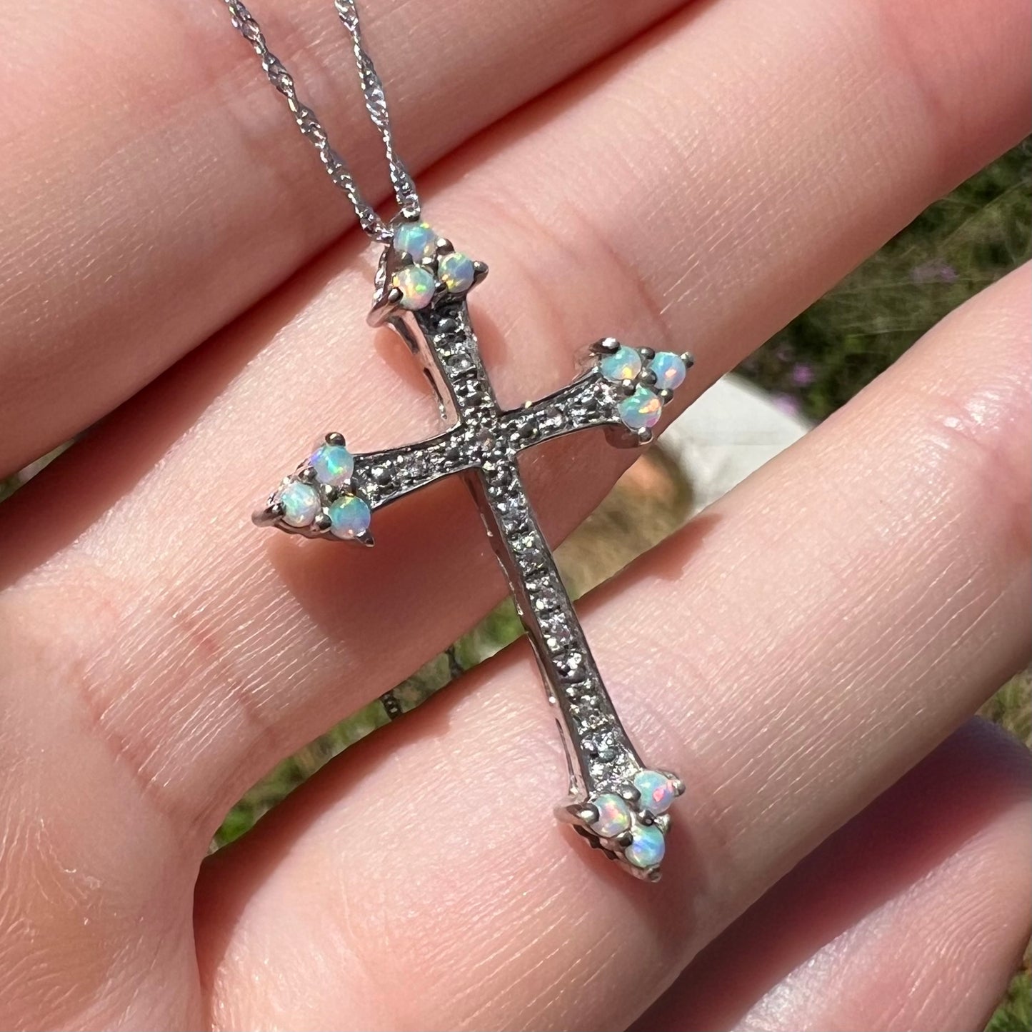 Sunlight view of an opal cross necklace held in a hand at a slight angle.