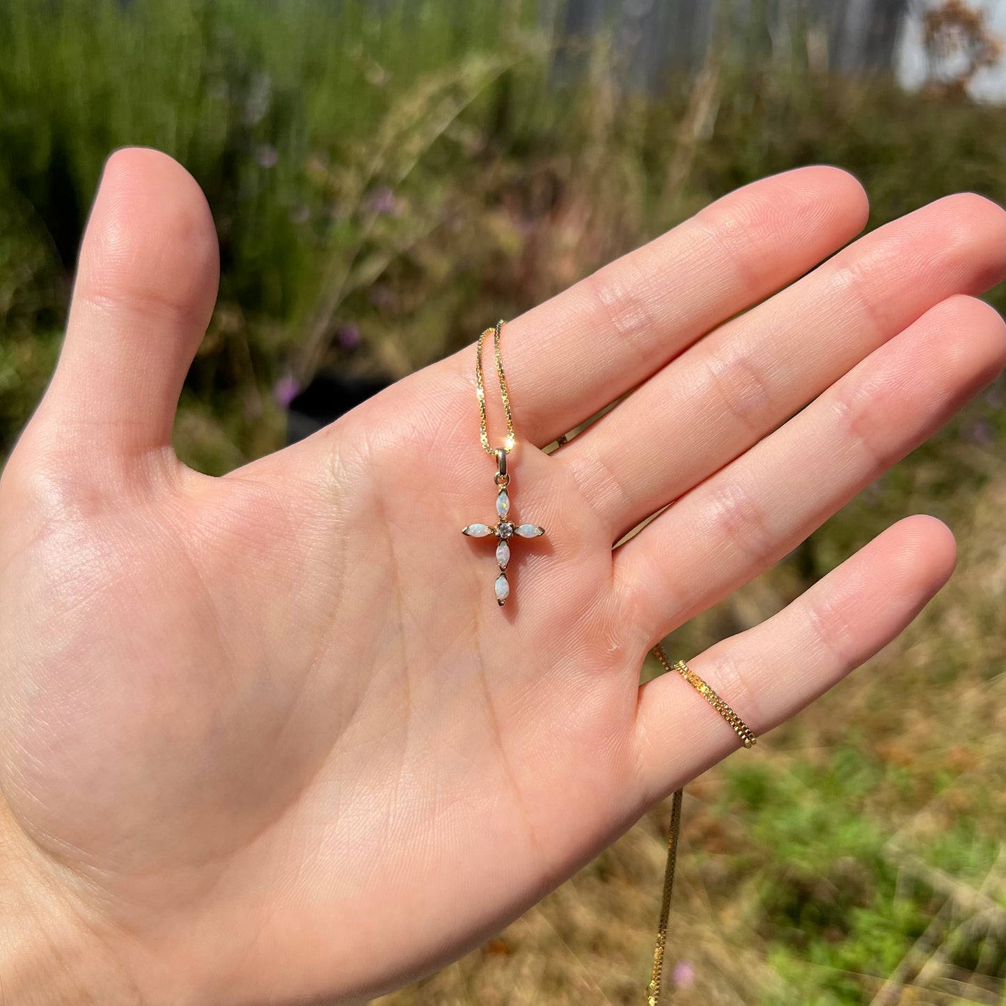 A dainty opal cross necklace shown at a distance, held in a hand in the sunlight.
