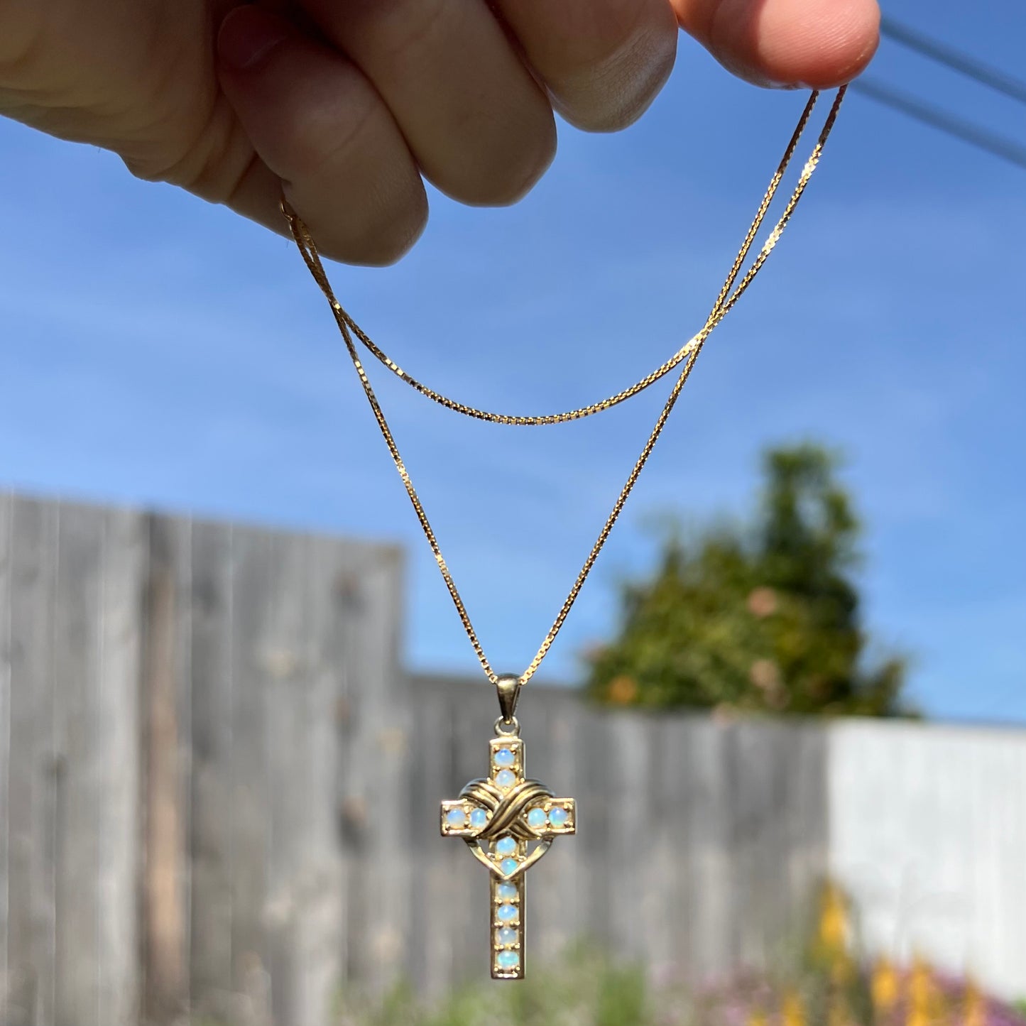 Opal cross necklace dangling in sunlight from hand.