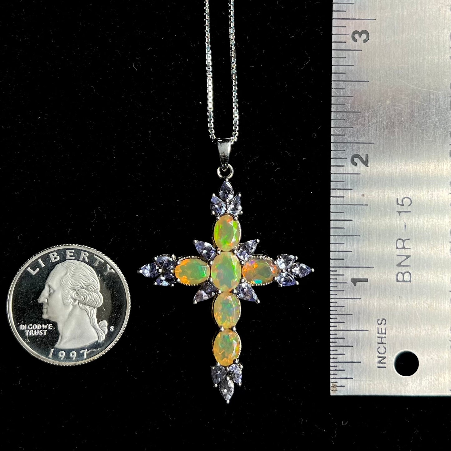 An opal cross necklace with that measures 2 inches long, laid next to a quarter and ruler.