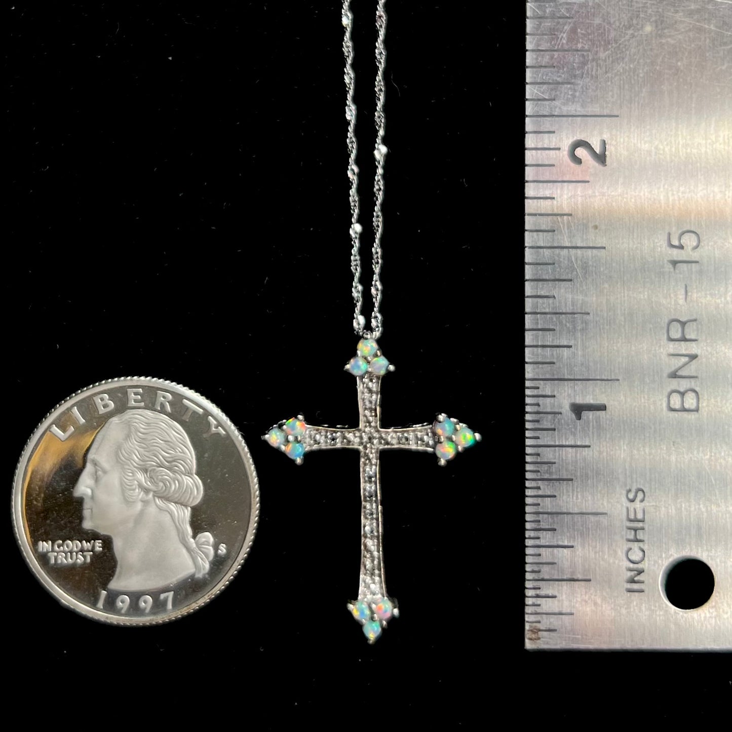 A silver opal cross necklace shown next to a quarter coin and ruler for scale.