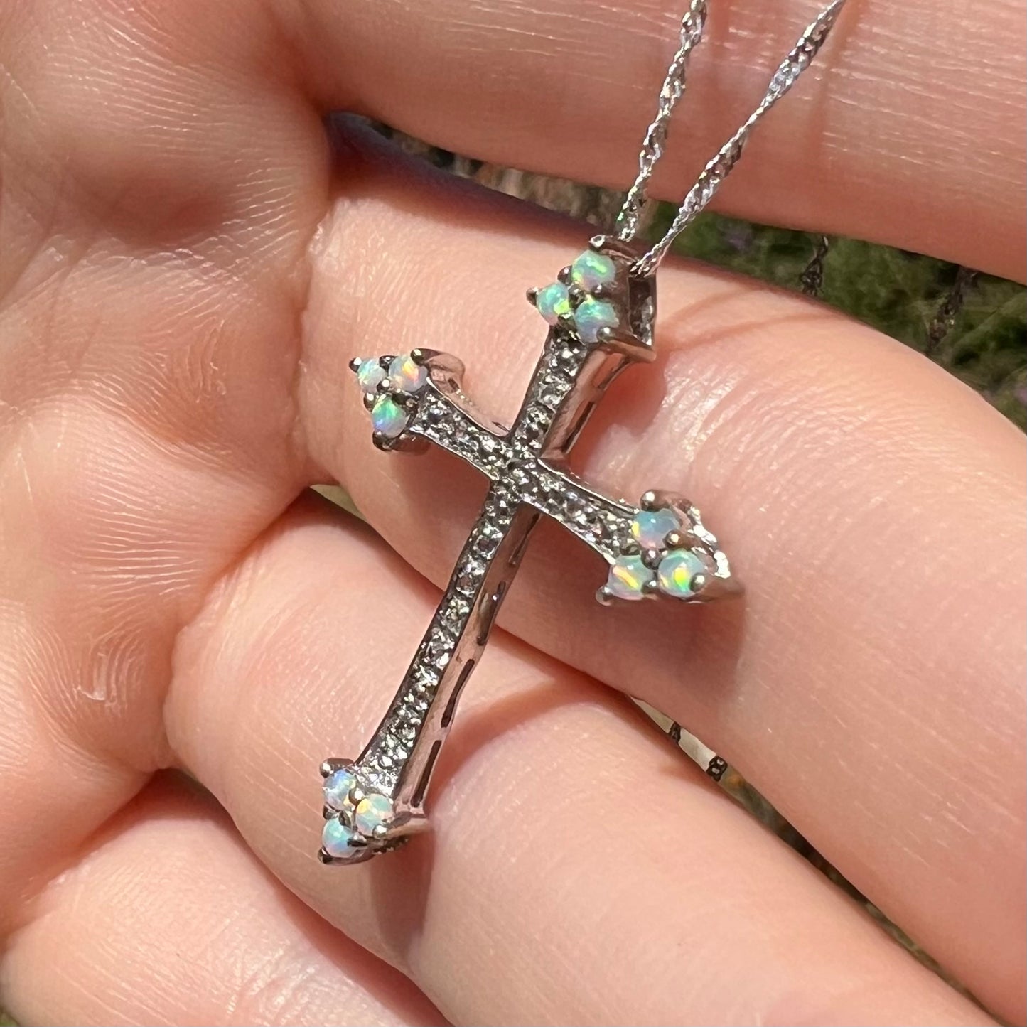 An opal cross necklace with CZ accents shown at an angle in natural light, held by a hand.