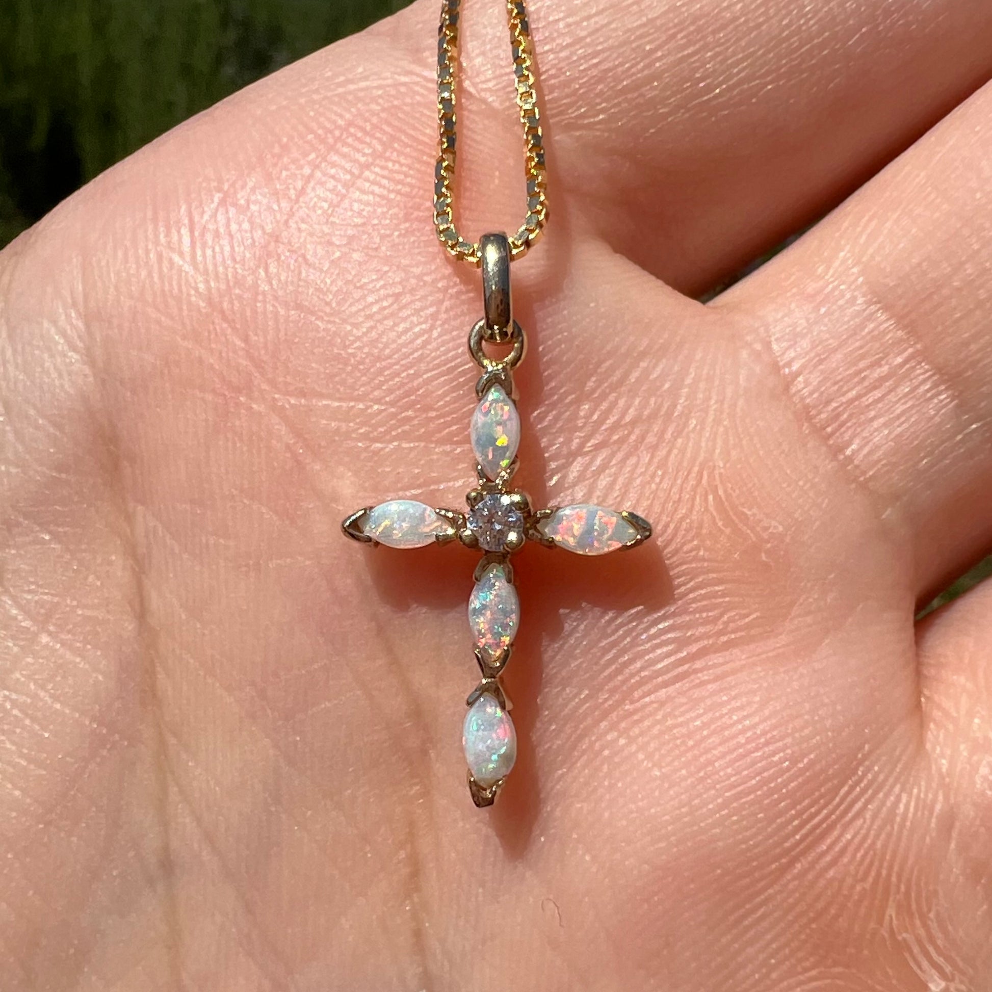 A dainty opal cross necklace shown in a hand, straight on, close-up in the sunlight.