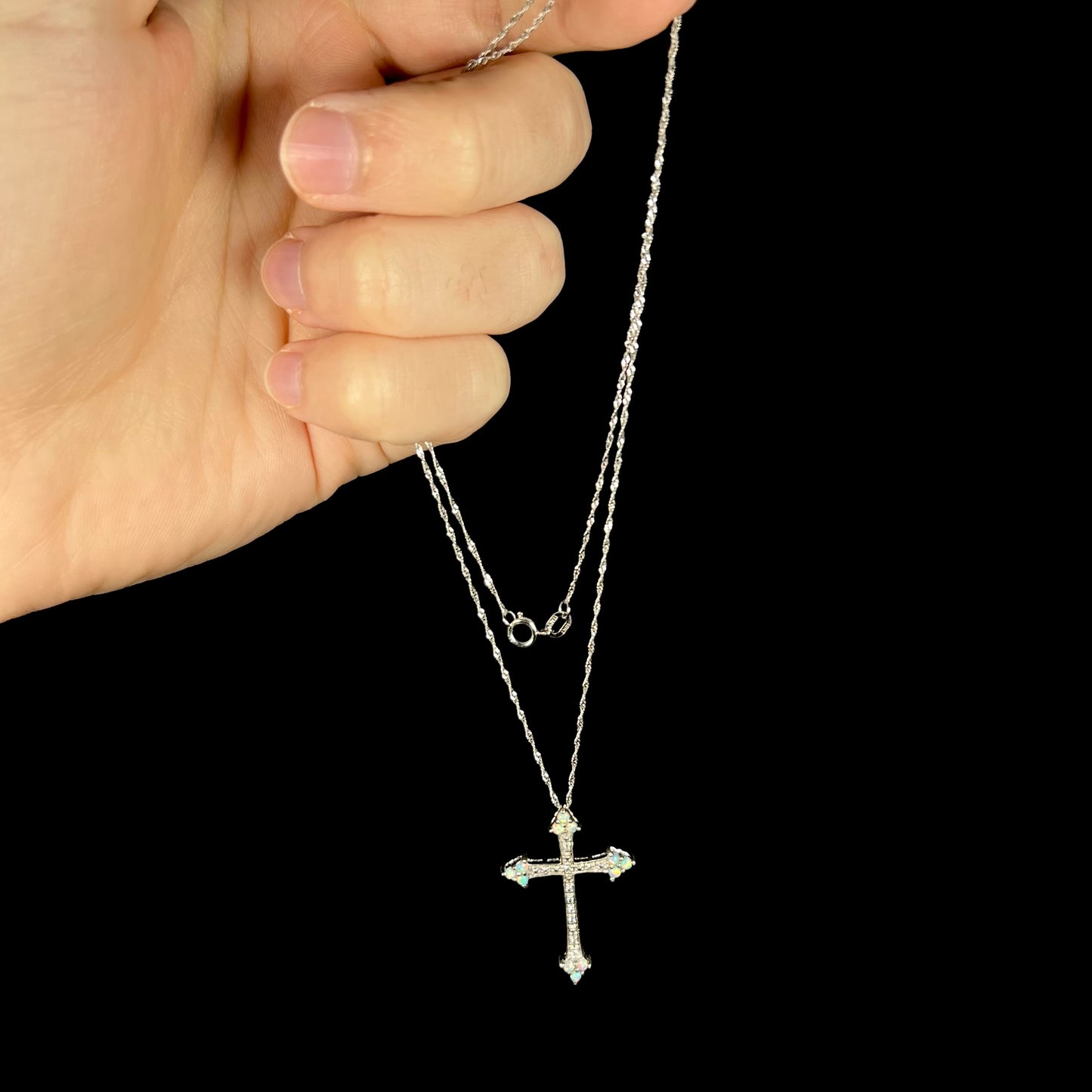 An opal cz cross necklace held in a hand from its silver chain against a black background.