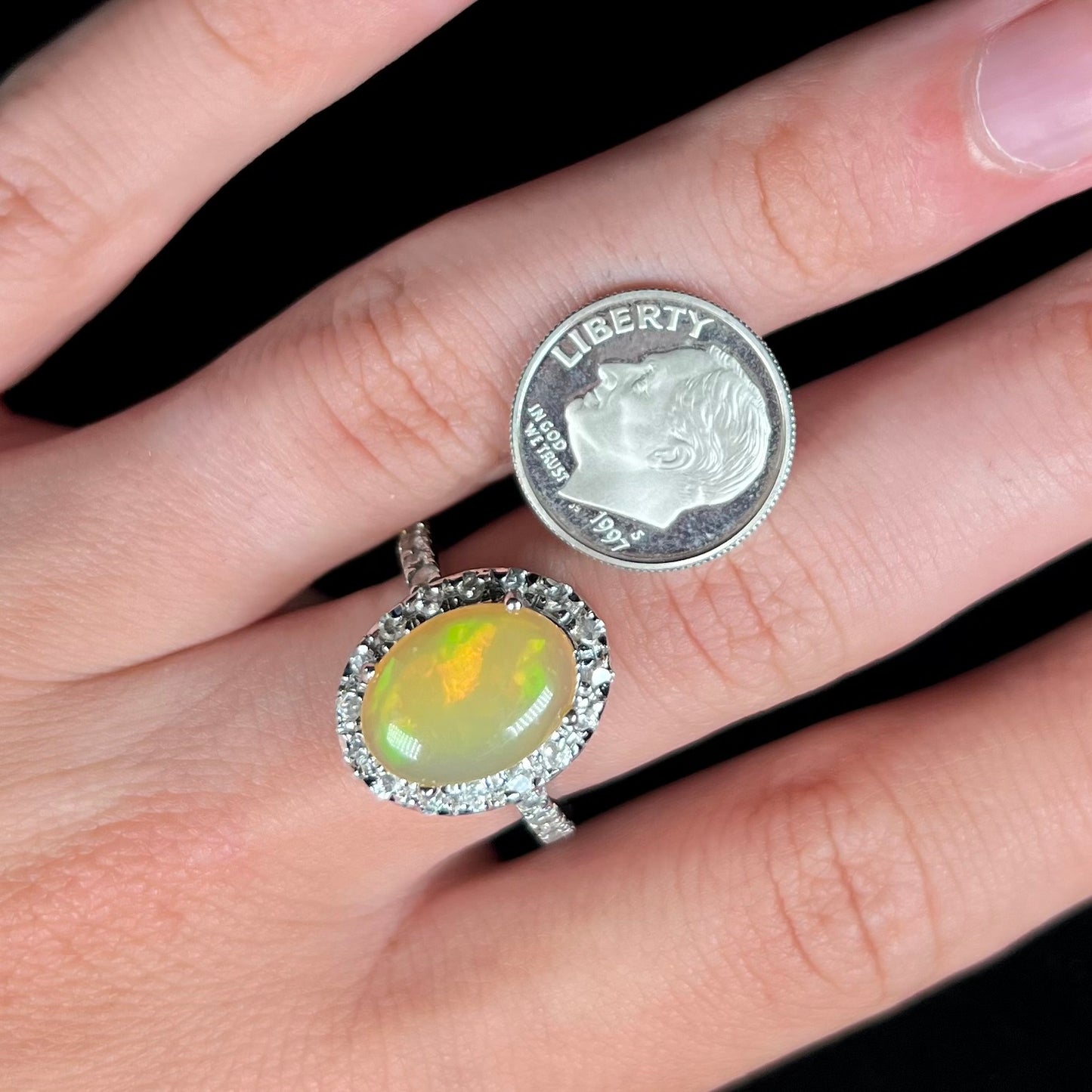 A sterling silver Ethiopian fire opal ring with a CZ halo worn on a hand next to a 1997 S dime coin.