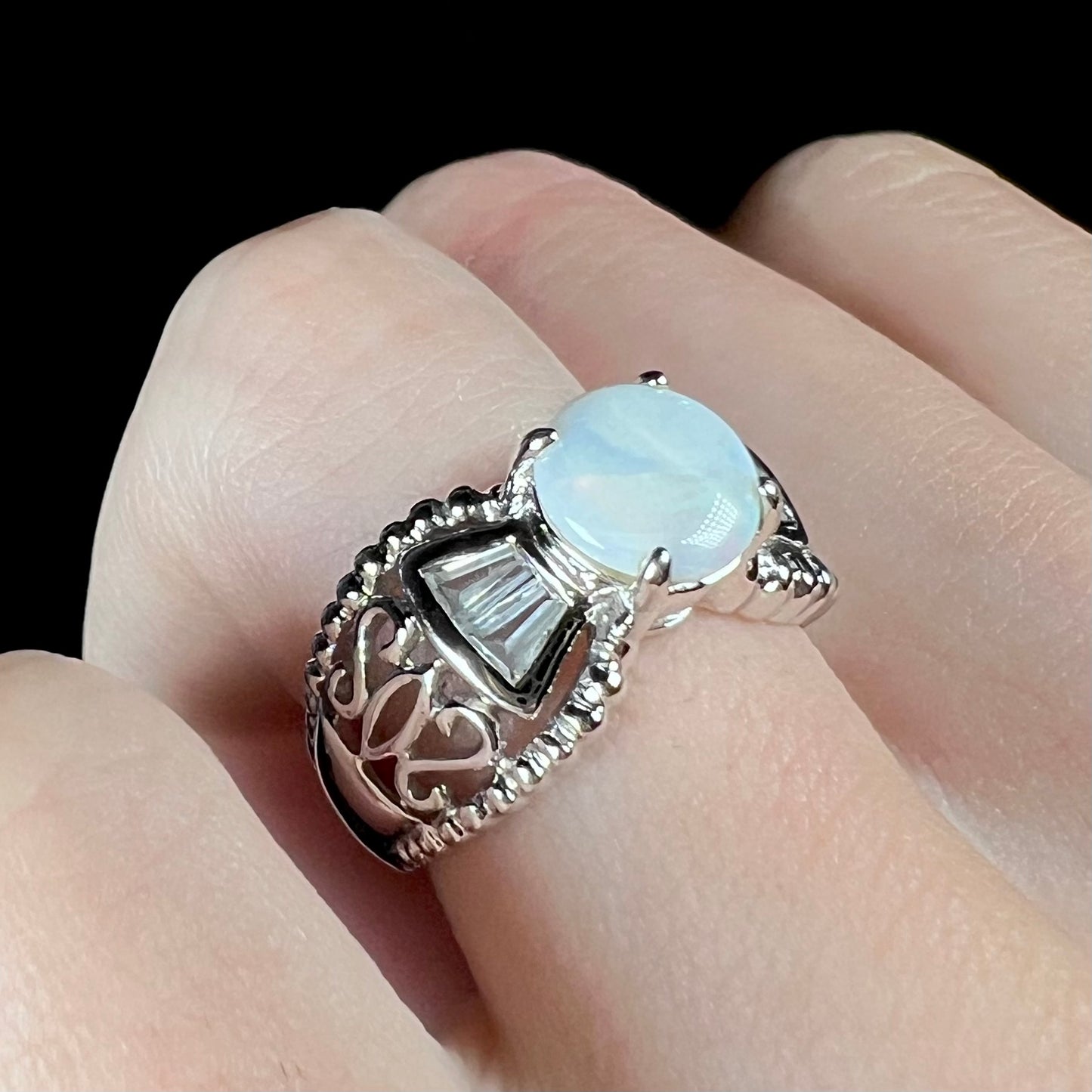 A round Australian white opal and CZ ring in white base metal worn on a finger and shown at an angle.