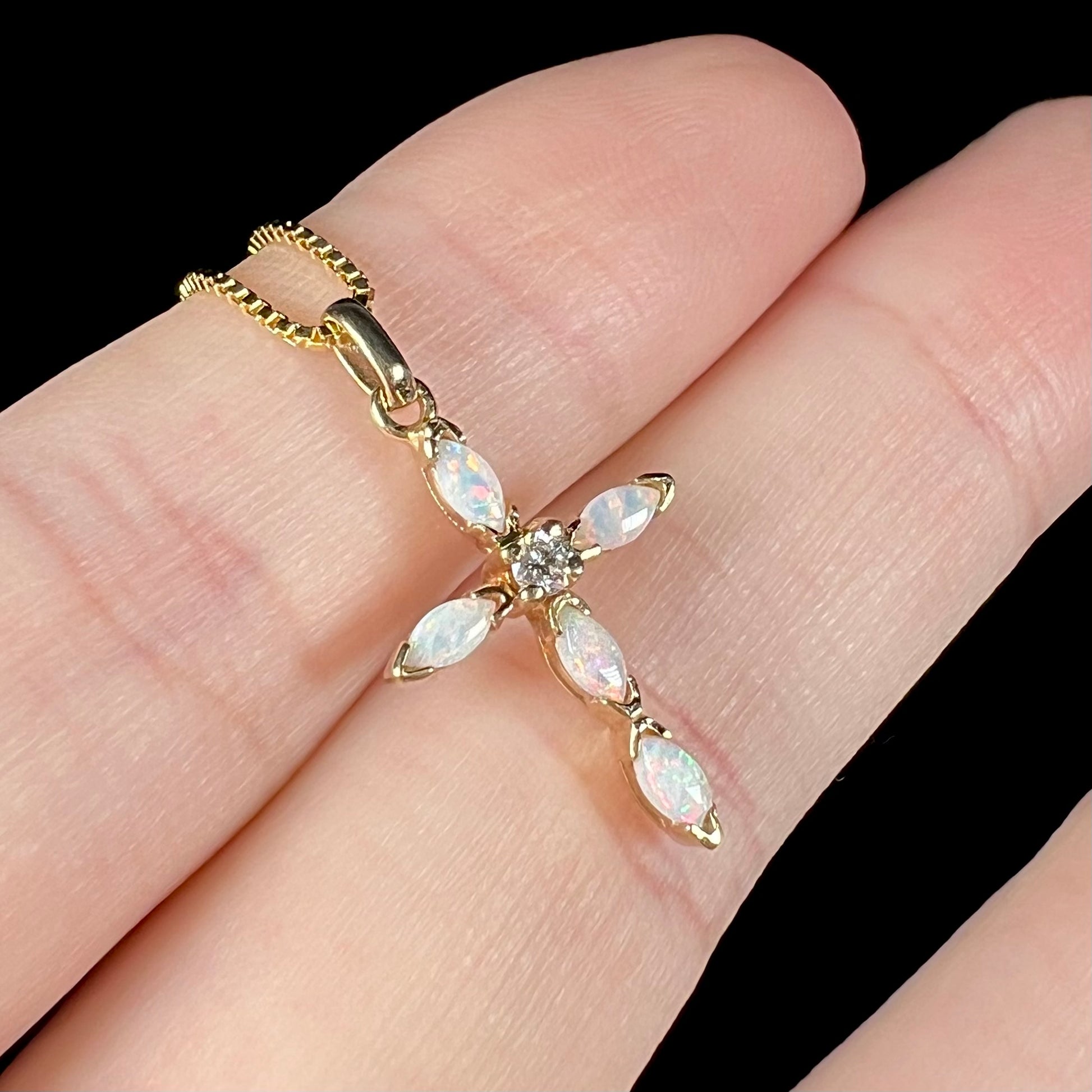 An opal and diamond cross necklace shown close-up at an angle, held by two fingers.