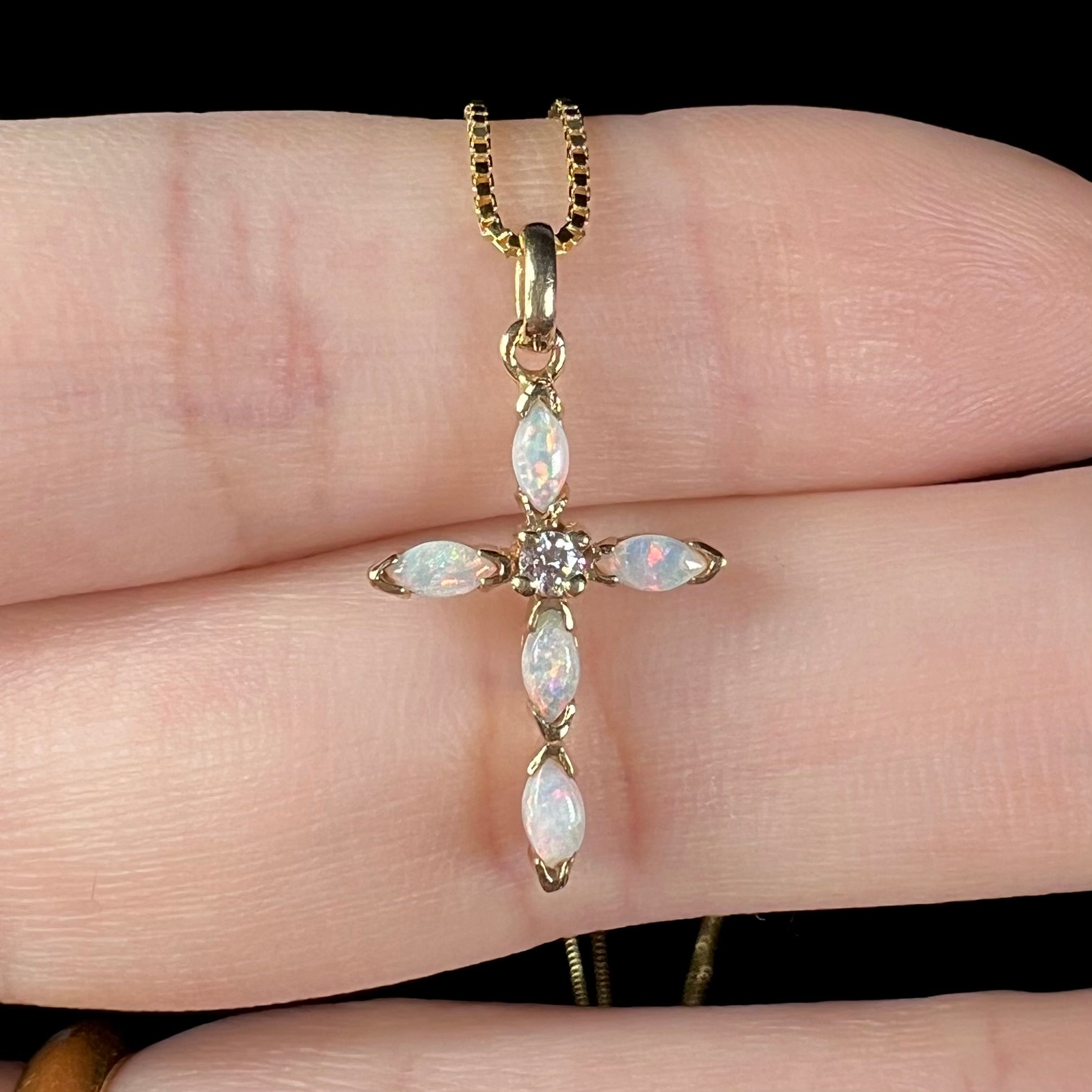 An opal and diamond cross necklace shown from the front close-up, held in a hand.