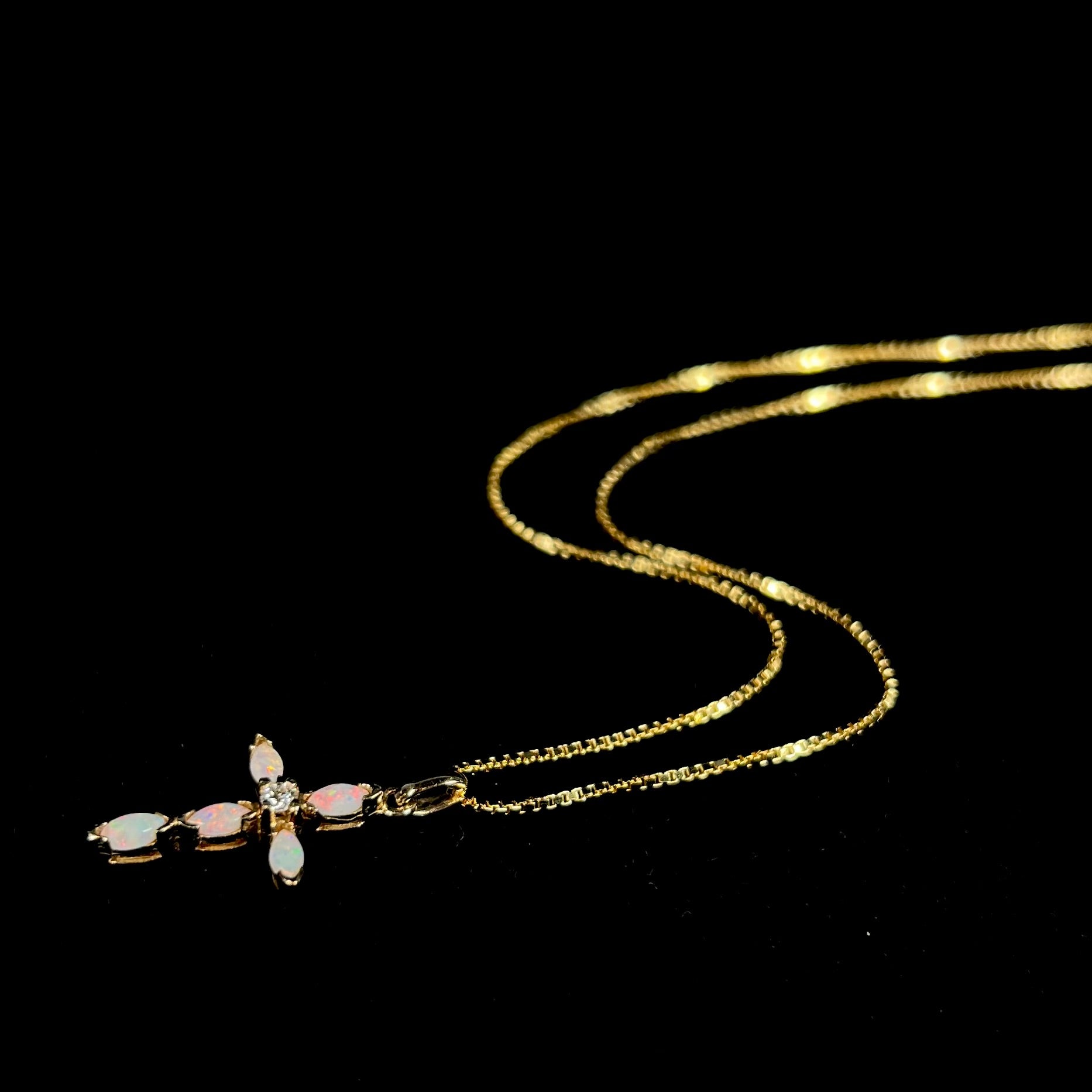 An opal and diamond cross necklace laying down with the chain splayed elegantly against black backdrop.