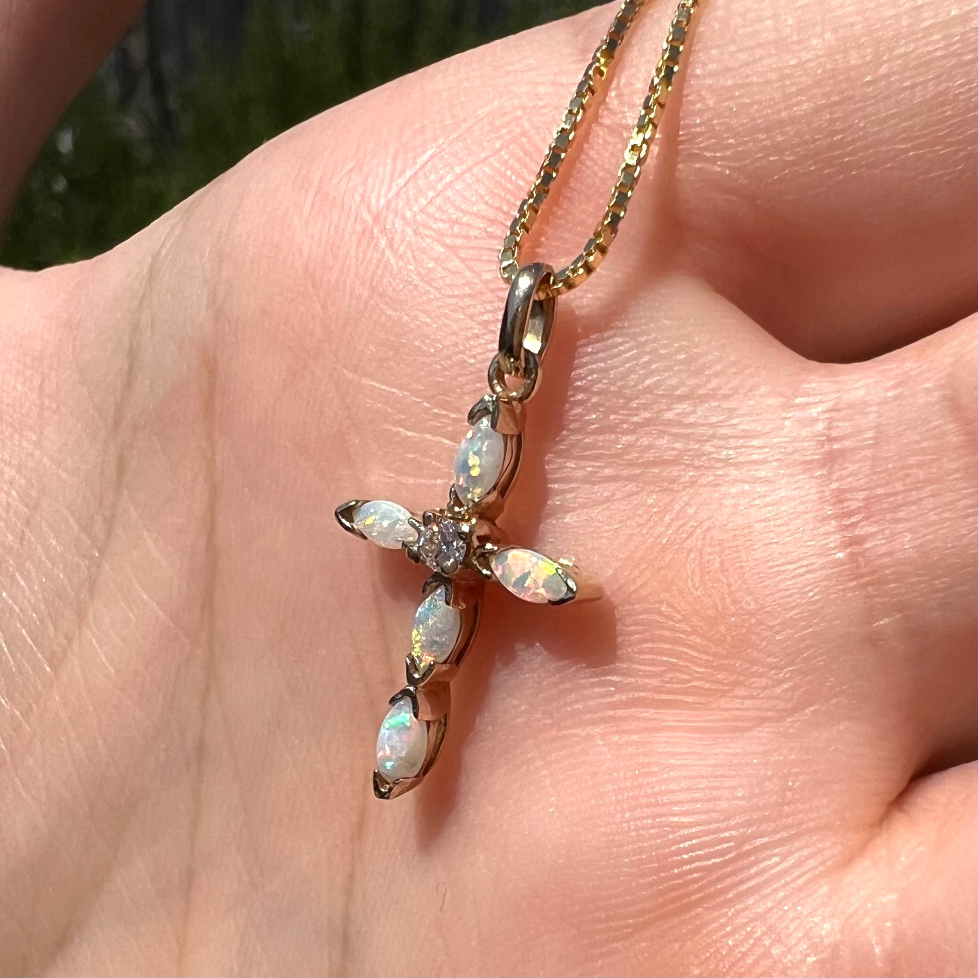 An opal diamond cross necklace shown in the sunlight, held in a hand at an angle.