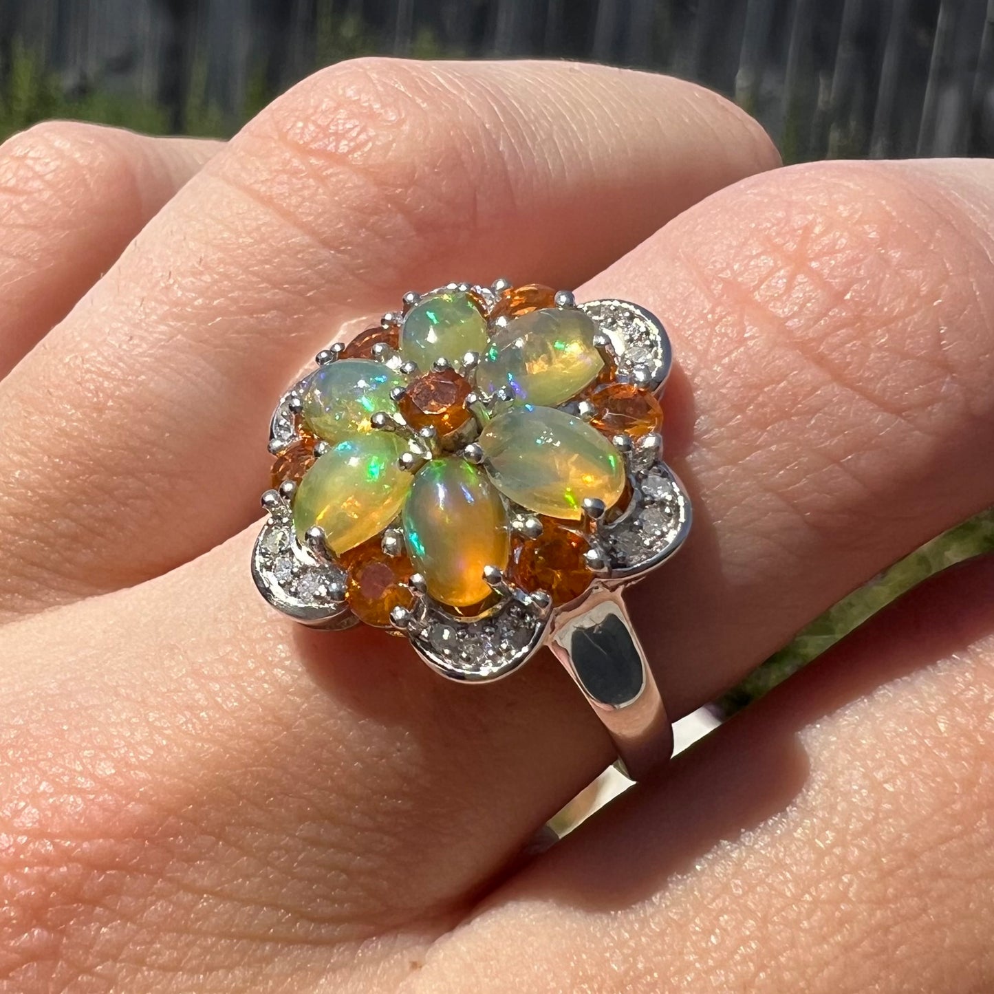 Angled view of an opal and diamond flower ring worn in the sunlight on a finger.