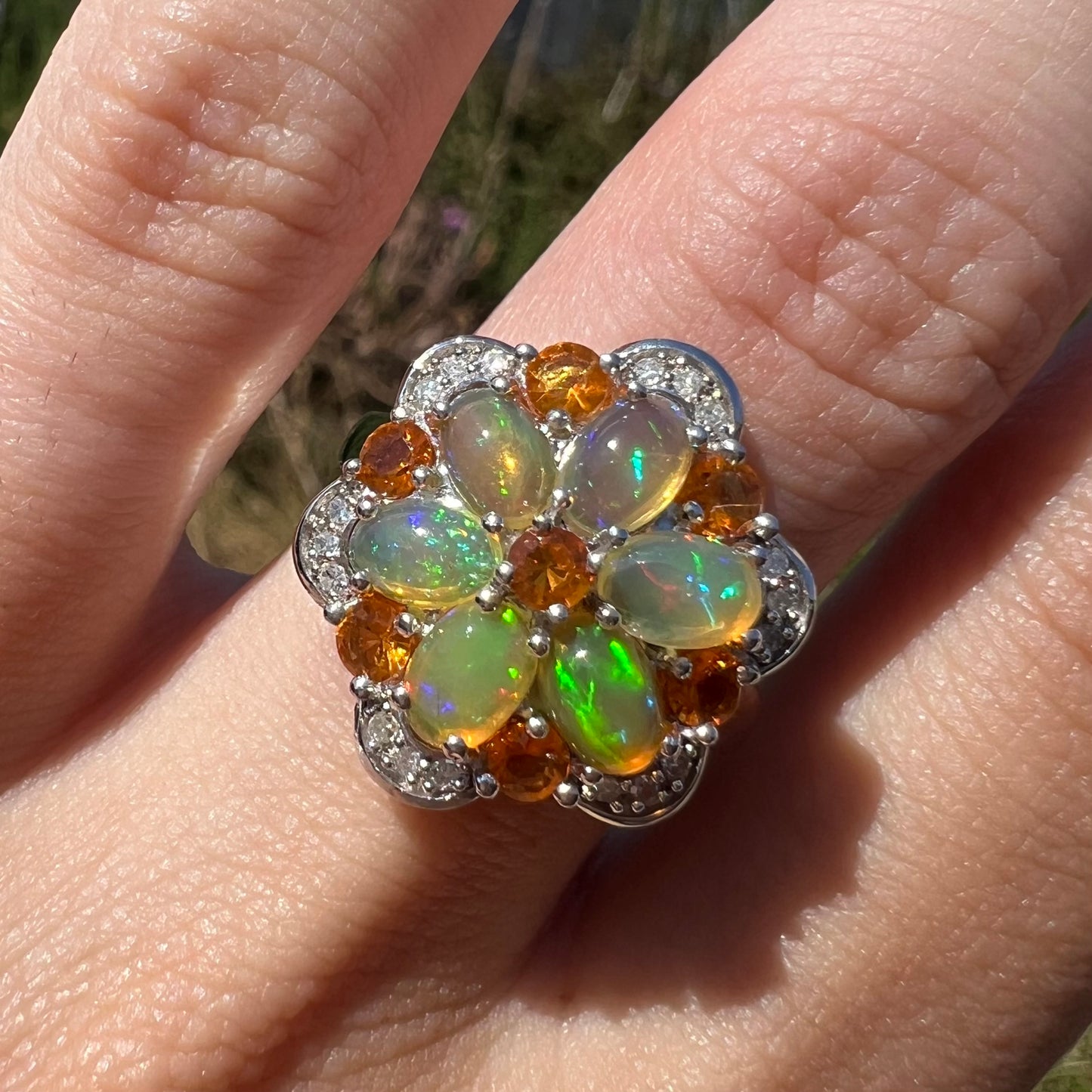 Front close-up of an opal and diamond flower ring in natural sunlight worn on a finger. The opals show green colors.
