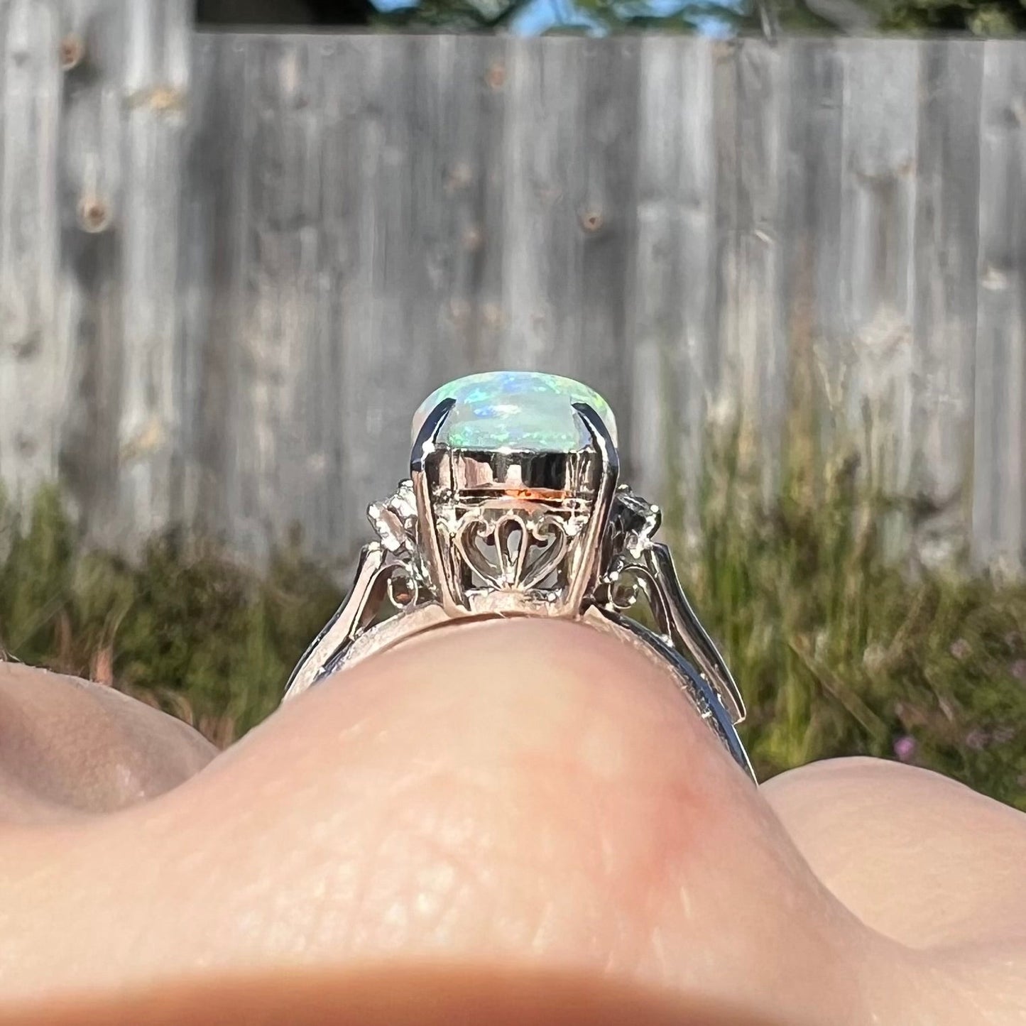 A platinum filigree opal engagement ring set with diamond accents. The opal has green-blue colors.