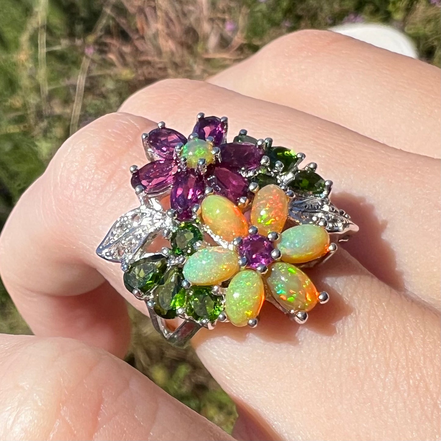 An opal flower ring worn on a finger at an angle outdoors in natural light, shown close-up.