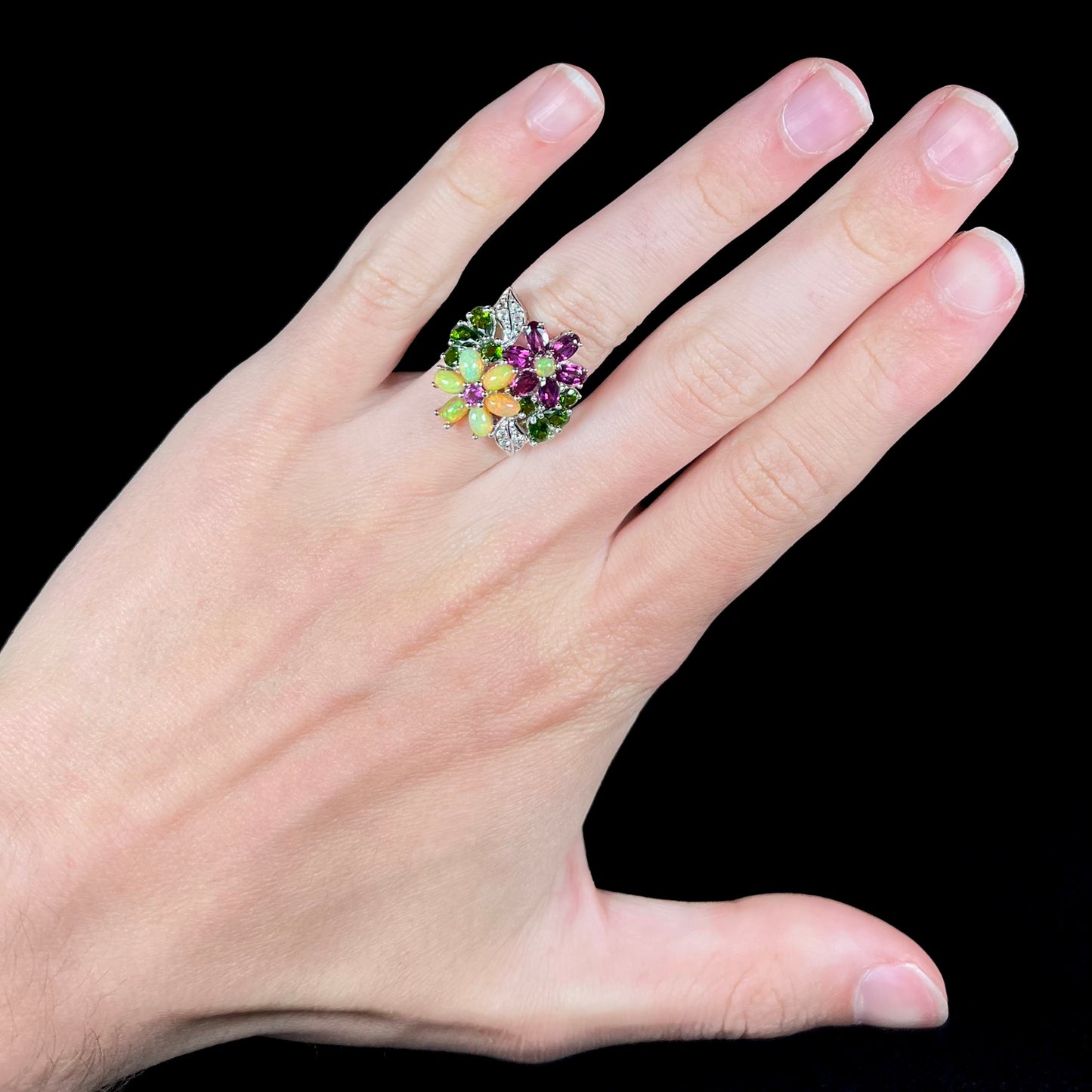 An opal and garnet flower ring, worn on a hand against a black background.