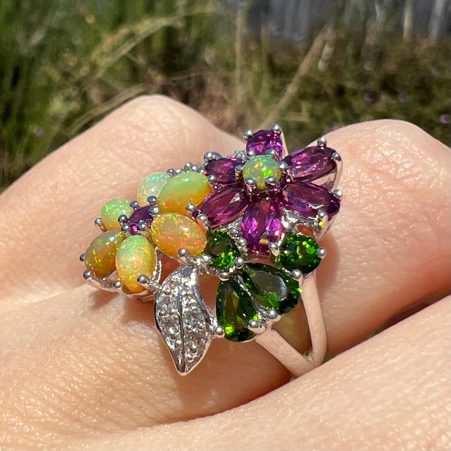 An opal and garnet gemstone flower ring, shown at an angle on a finger close-up in the sunlight.