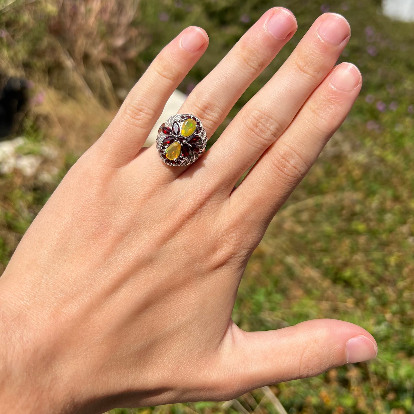 A sterling silver Ethiopian opal and garnet infinity-design ring with diamond accents, worn on a hand in sunlight.
