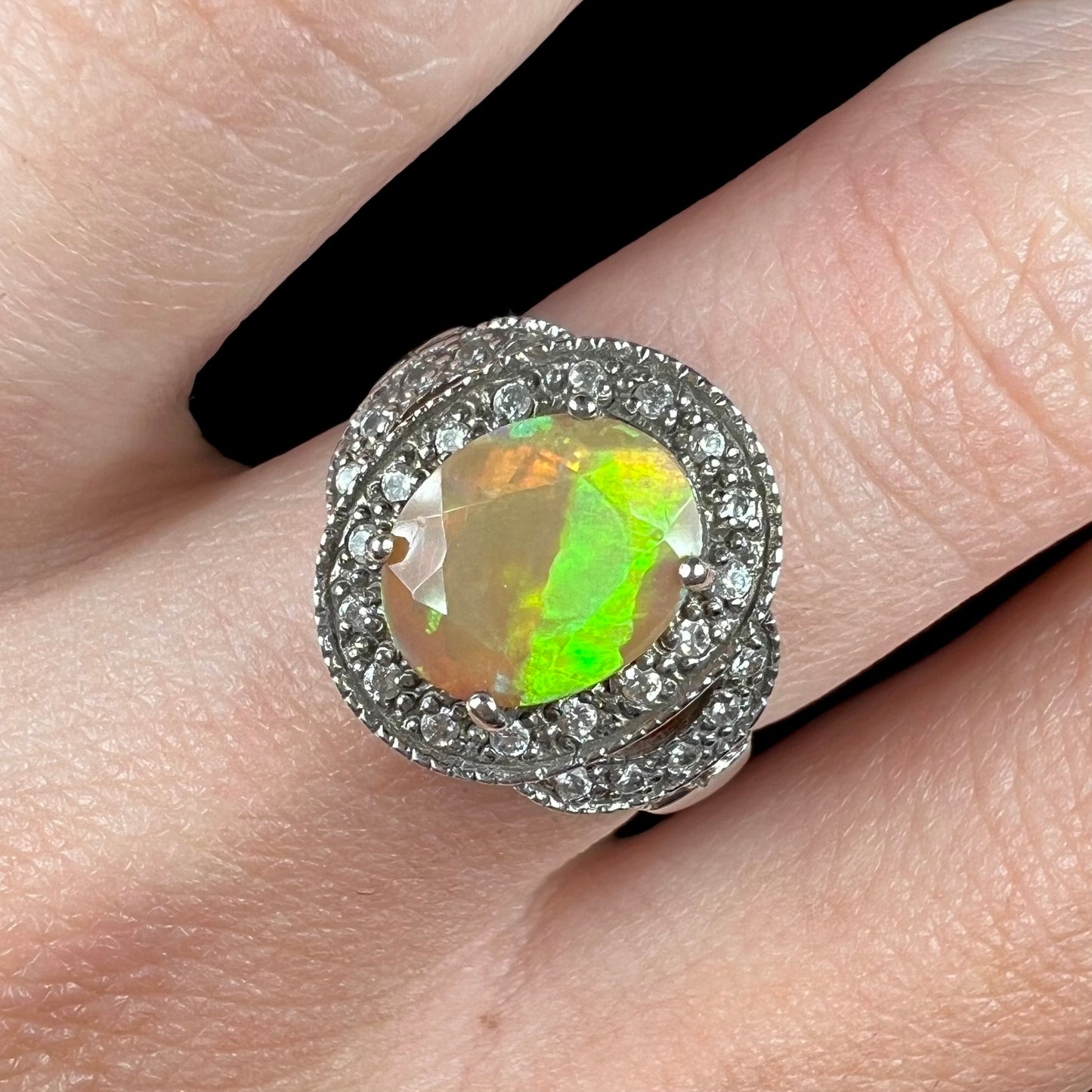 Macro close-up of an opal halo ring worn on a finger. The opal shows a section of green color play.