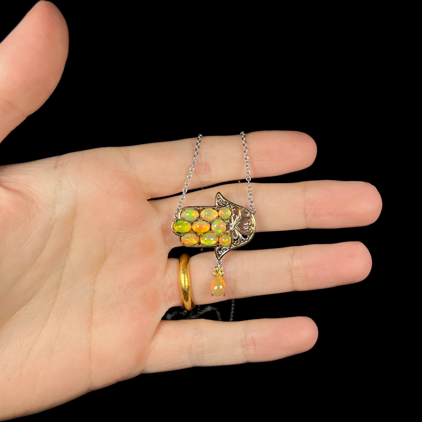 An opal hamsa necklace with Ethiopian fire opals, held in a hand against a black background.