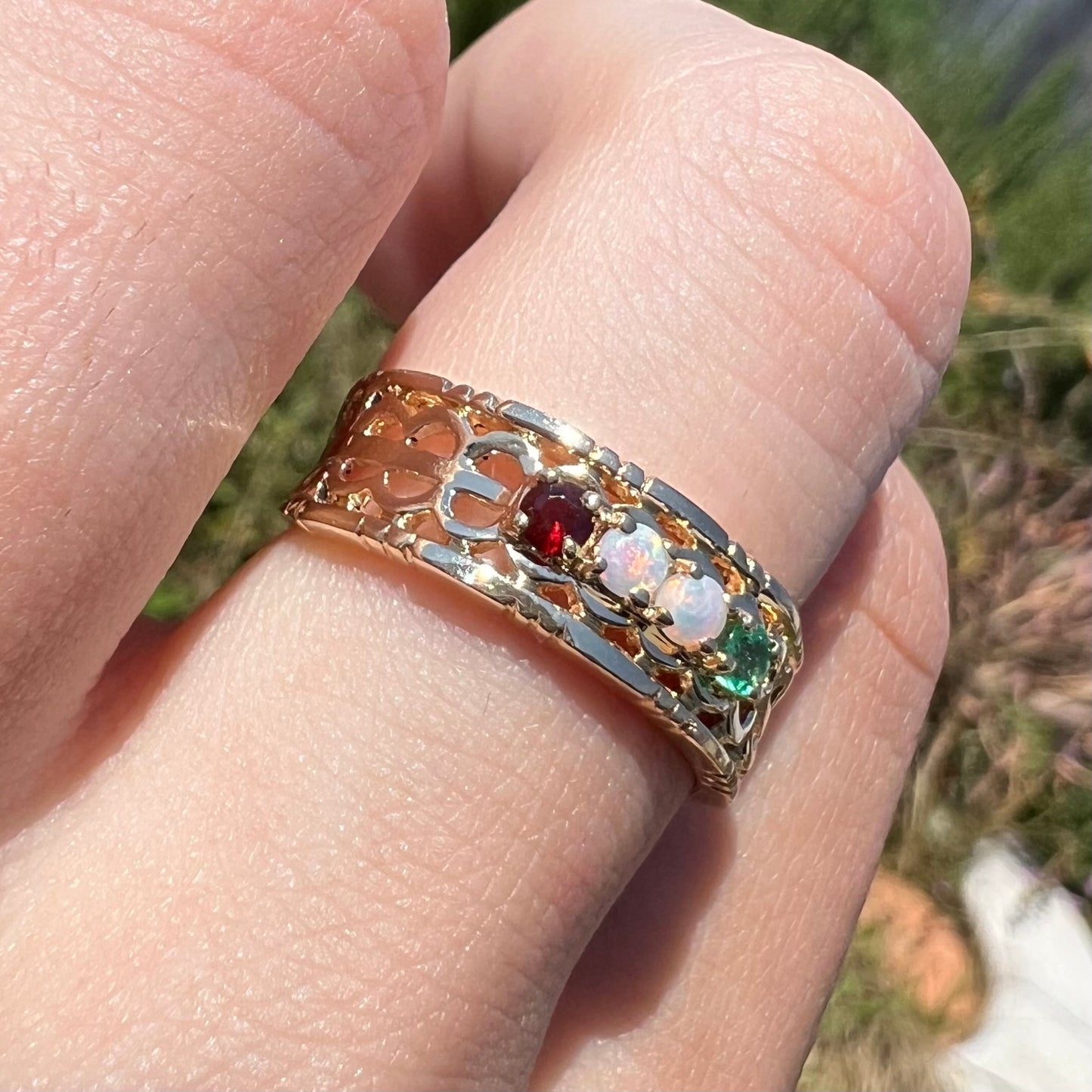 A gold open-design mother's ring set with two opals, an emerald, and a garnet, worn in the sunlight.
