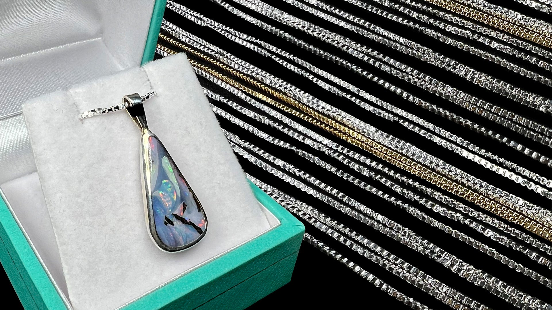 A pear shaped Australian boulder opal pendant shown in a display box next to a row of sterling silver box chains.