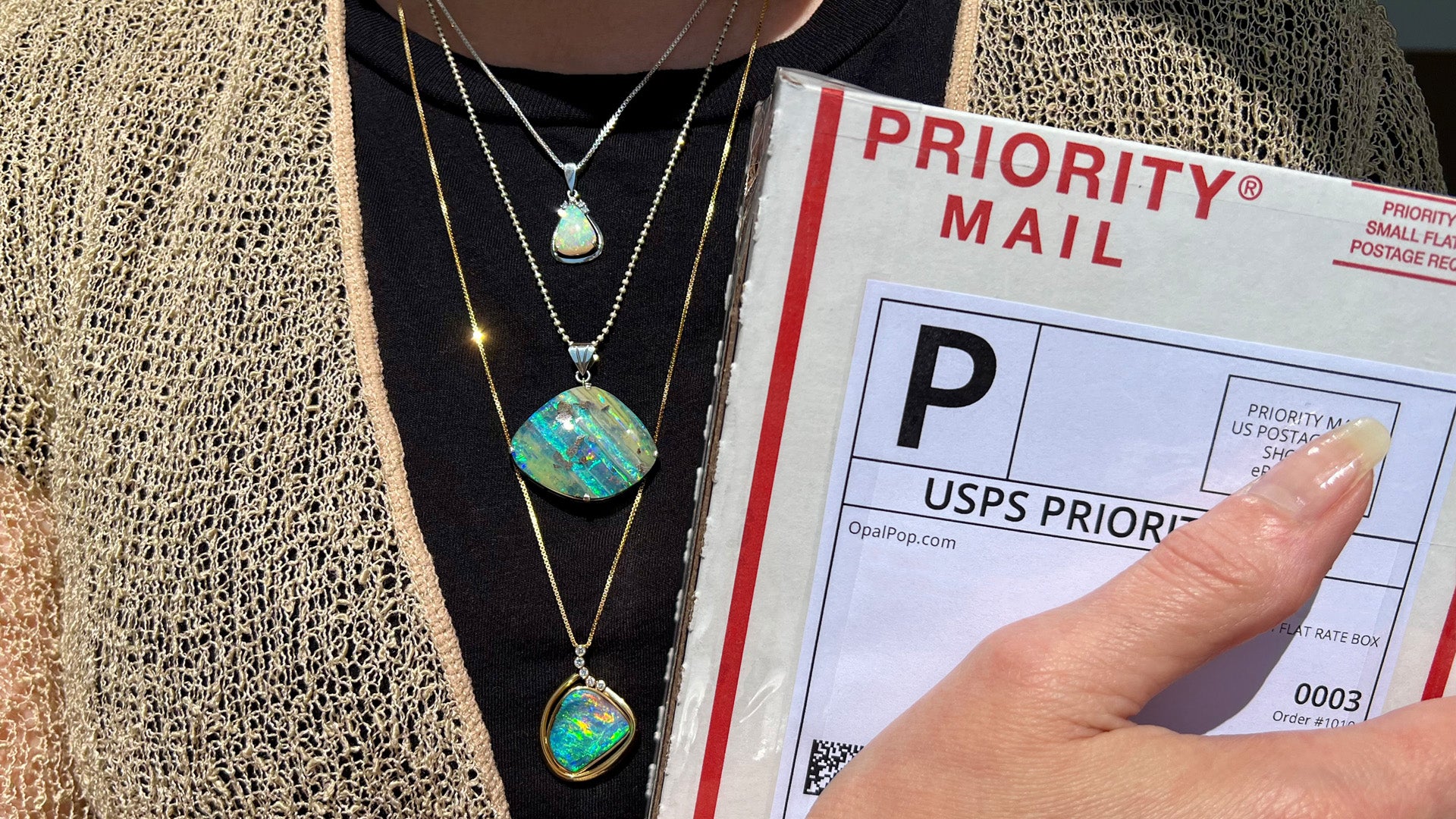 A woman holding a USPS small flat rate box to her chest, wearing three layered opal necklaces of different lengths.