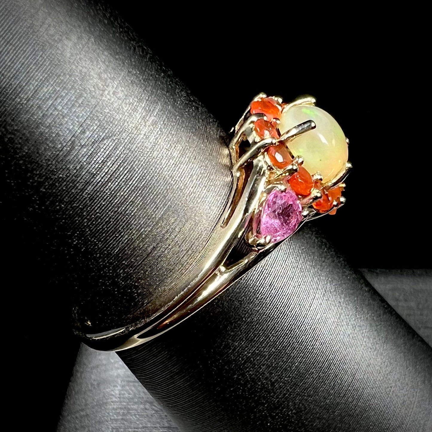 A yellow gold ring mounted with a round Ethiopian opal in a halo of faceted Mexican fire opals. Two pear shaped pink tourmalines accent the sides of the ring.