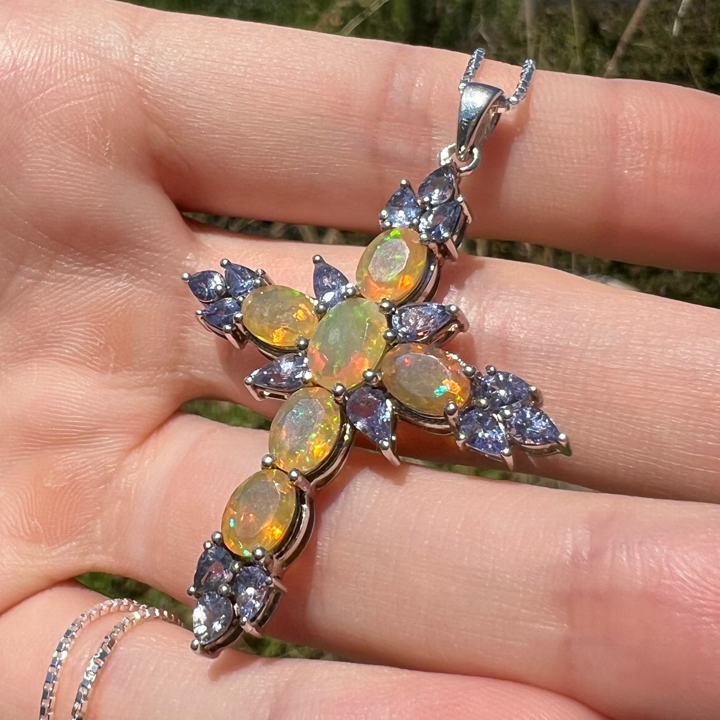 A silver opal and tanzanite cross necklace, held in the sunlight by a hand at an angle.