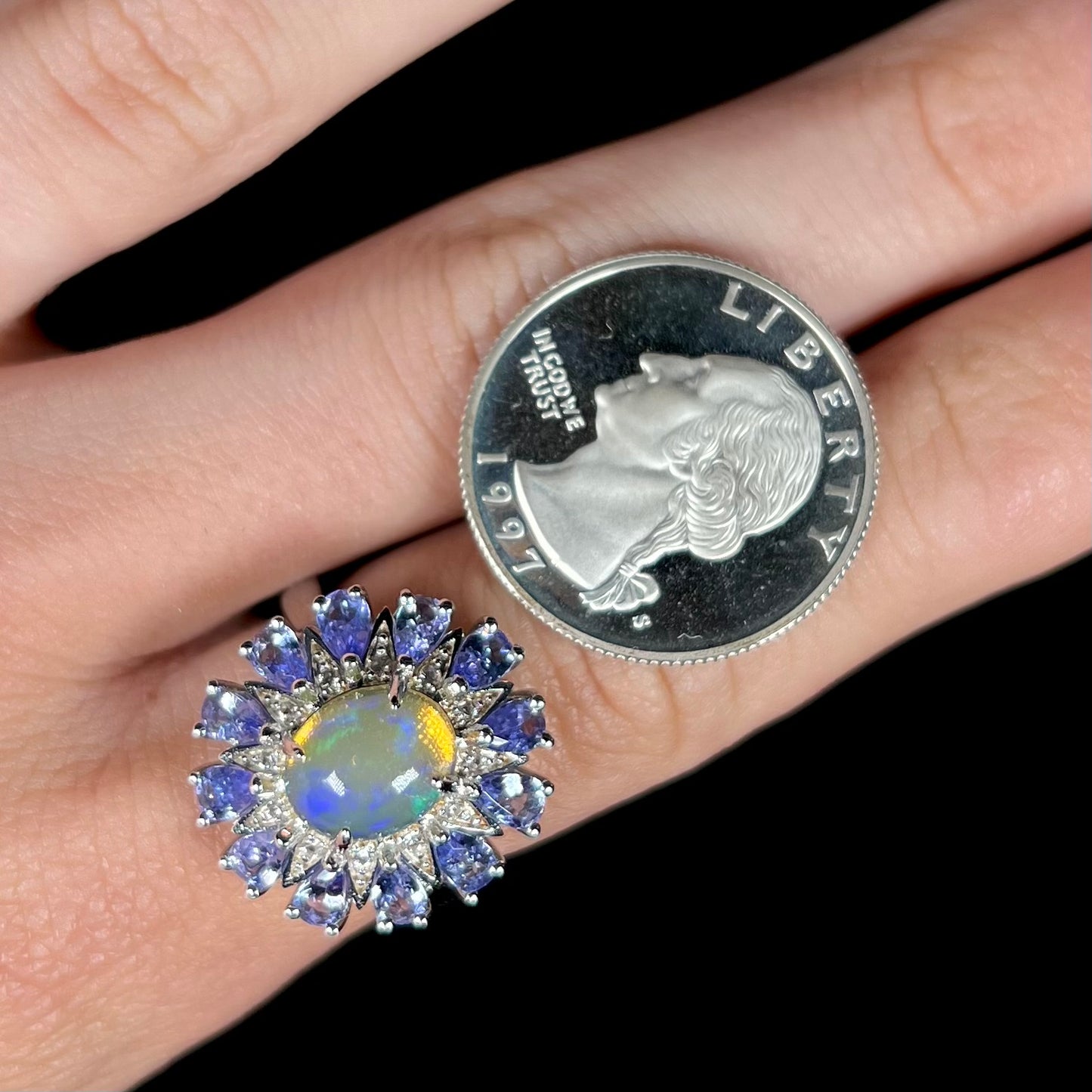 An opal and tanzanite halo ring worn on a finger next to a quarter coin for scale.