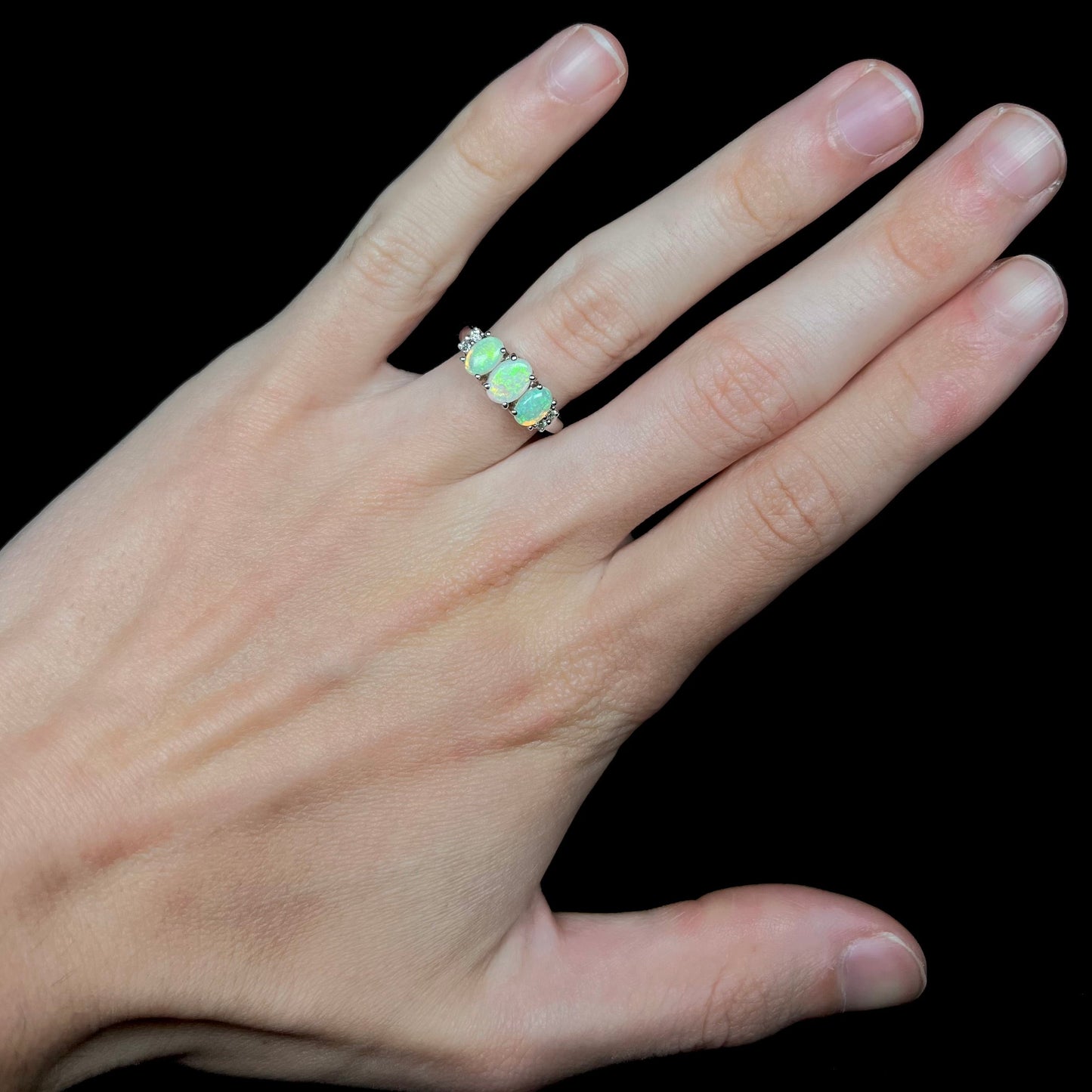 A ladies' white gold ring prong-set with three natural white opals and diamond accents. The opals play a green color.