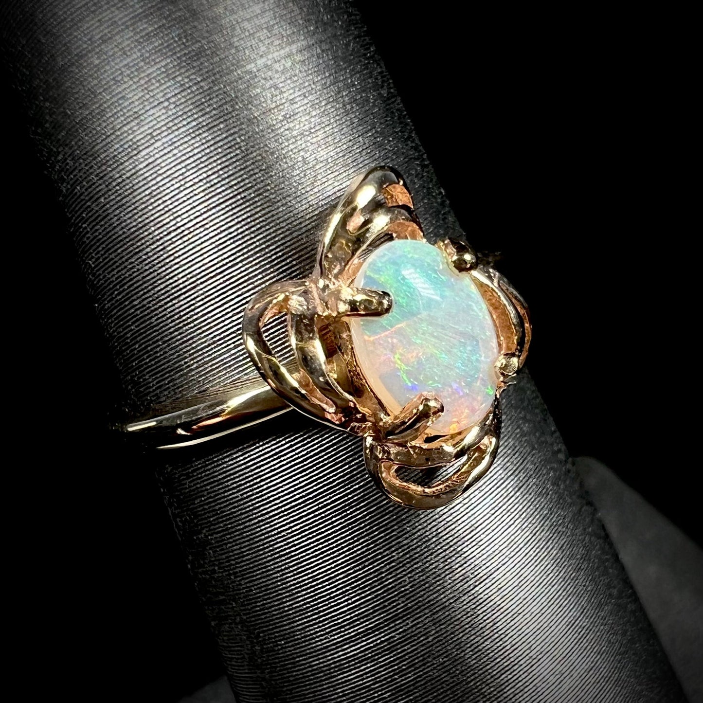 A yellow gold ring mounted with an oval cut opal. The opal has a white base tone with green and blue color play.