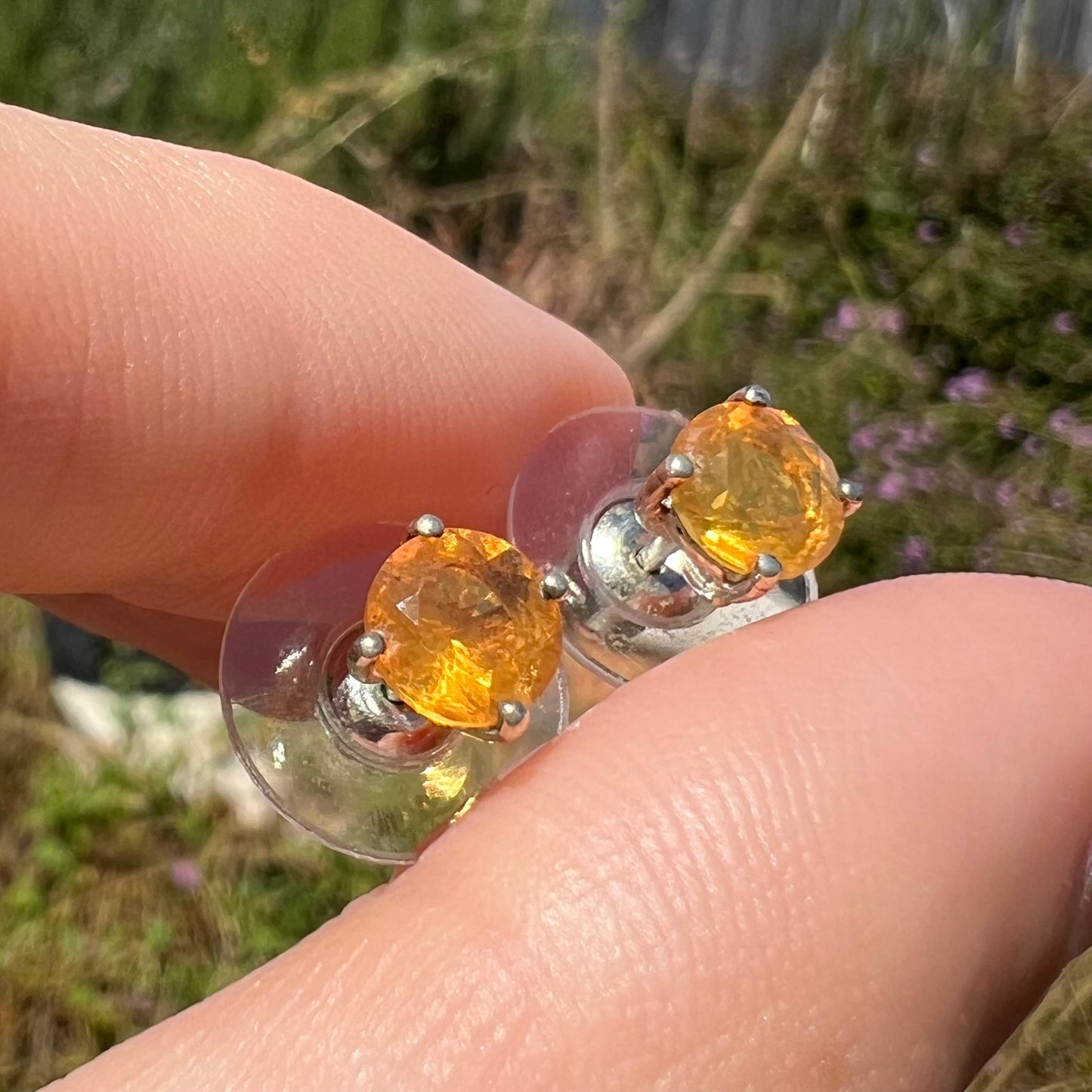 A pair of Mexican fire opal stud earrings in sterling silver, held an an angle in sunlight.