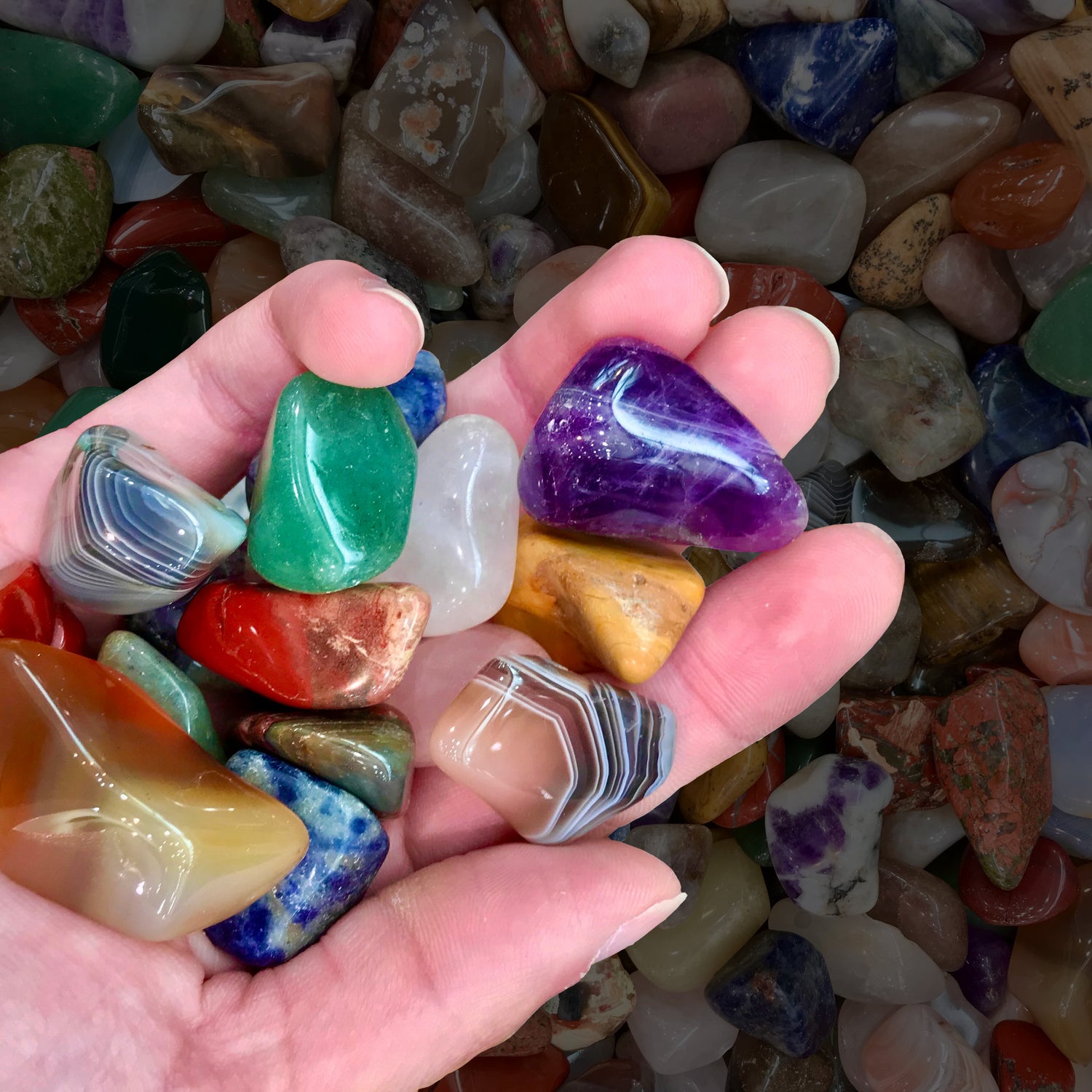 Large tumble polished stones are being held in a person's hand. Varieties seen include amethyst, agates, aventurine, and sodalite.