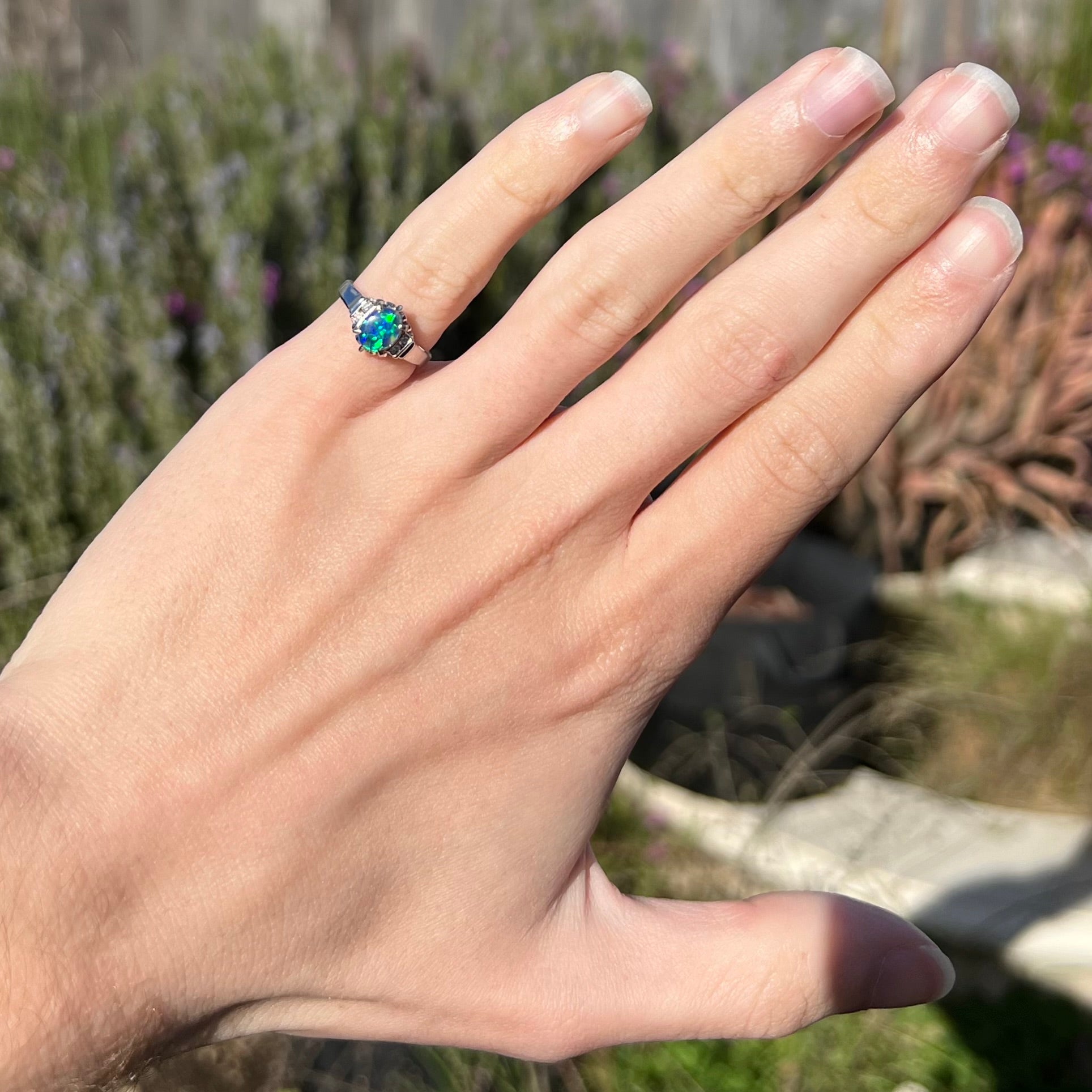 A petite black opal engagement ring is worn on the pinkie finger of a hand in natural sunlight.  The ring is platinum and has diamond accents.