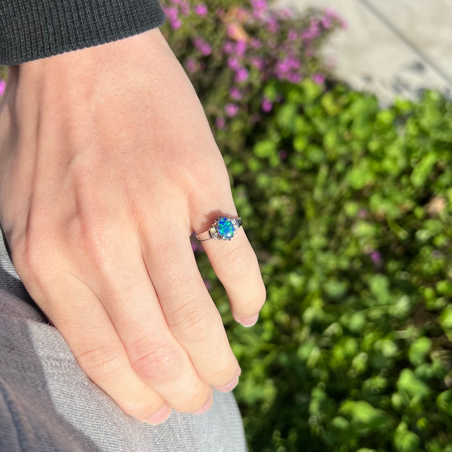 A petite platinum black opal and diamond engagement ring worn on a hand in the sunlight.  The opal displays blue color play.