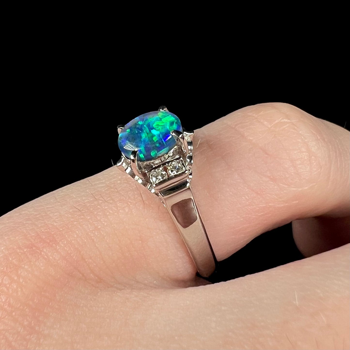 A pinkie finger wearing a petite platinum black opal engagement ring with diamond accents.  The opal shows brilliant green color play.
