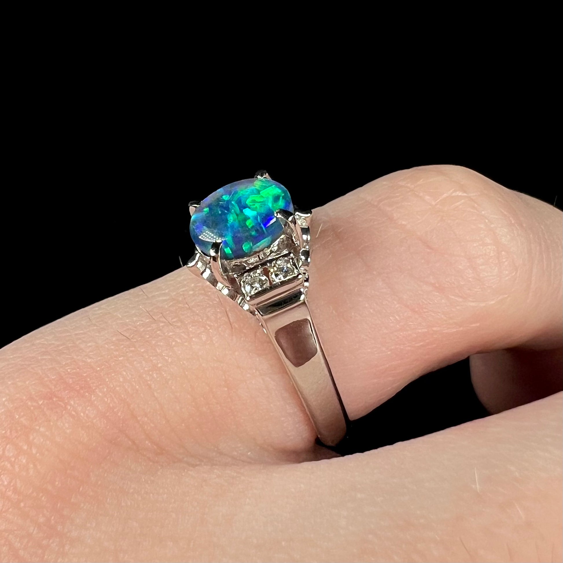 A pinkie finger wearing a petite platinum black opal engagement ring with diamond accents.  The opal shows brilliant green color play.