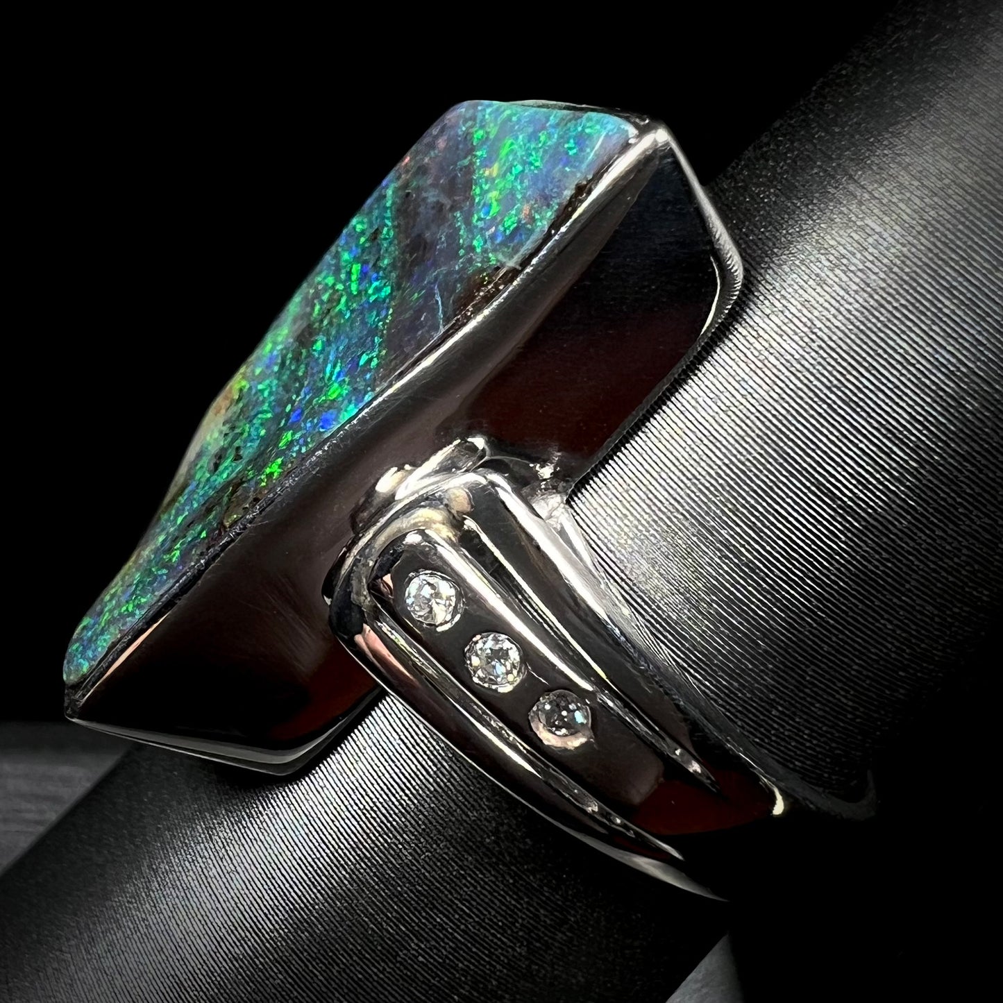 A platinum boulder opal ring with three visible diamond accents, shown from the side view on a ring display.